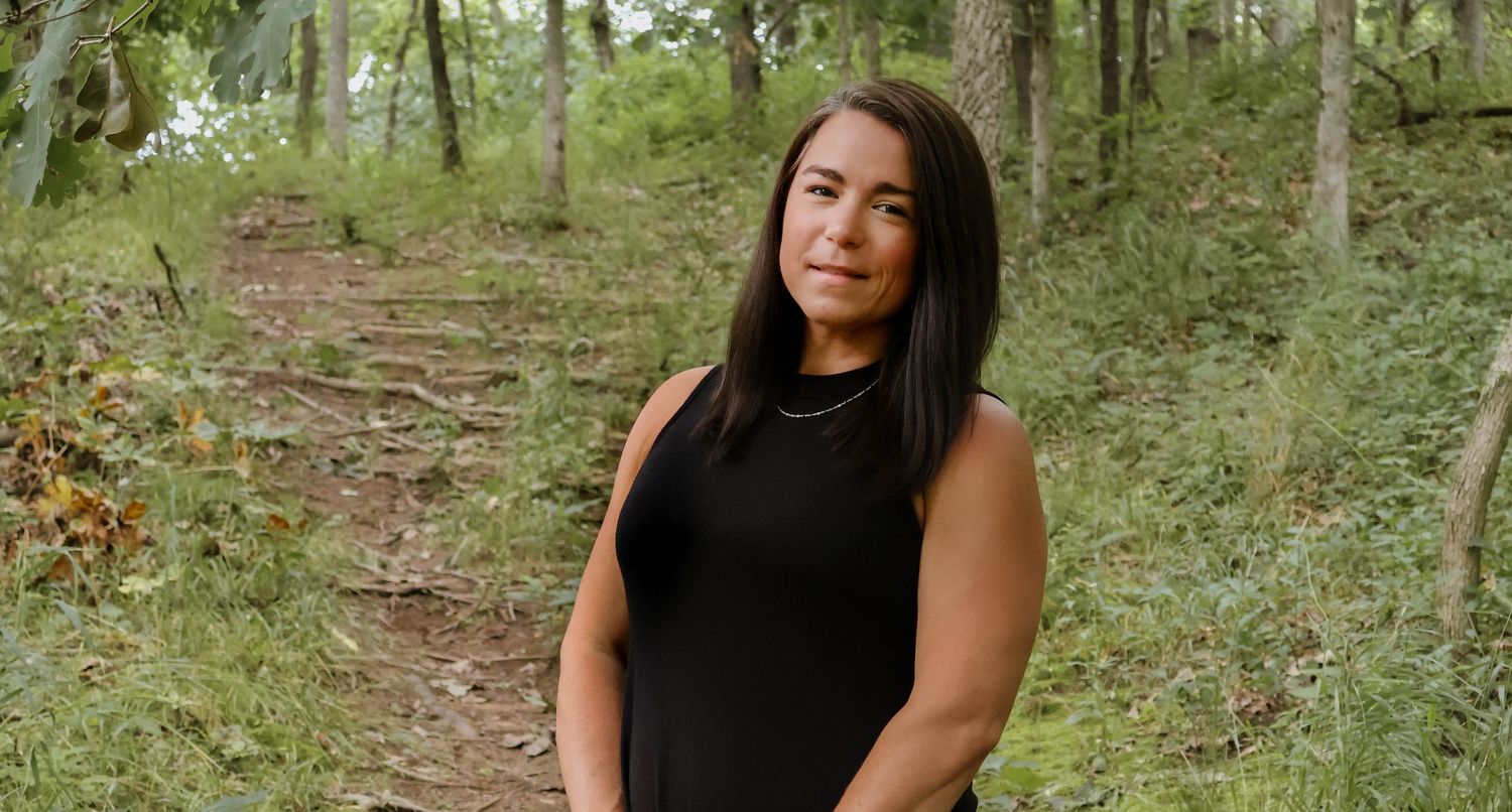 A person in a black sleeveless top stands confidently on a forest path surrounded by green trees and foliage.