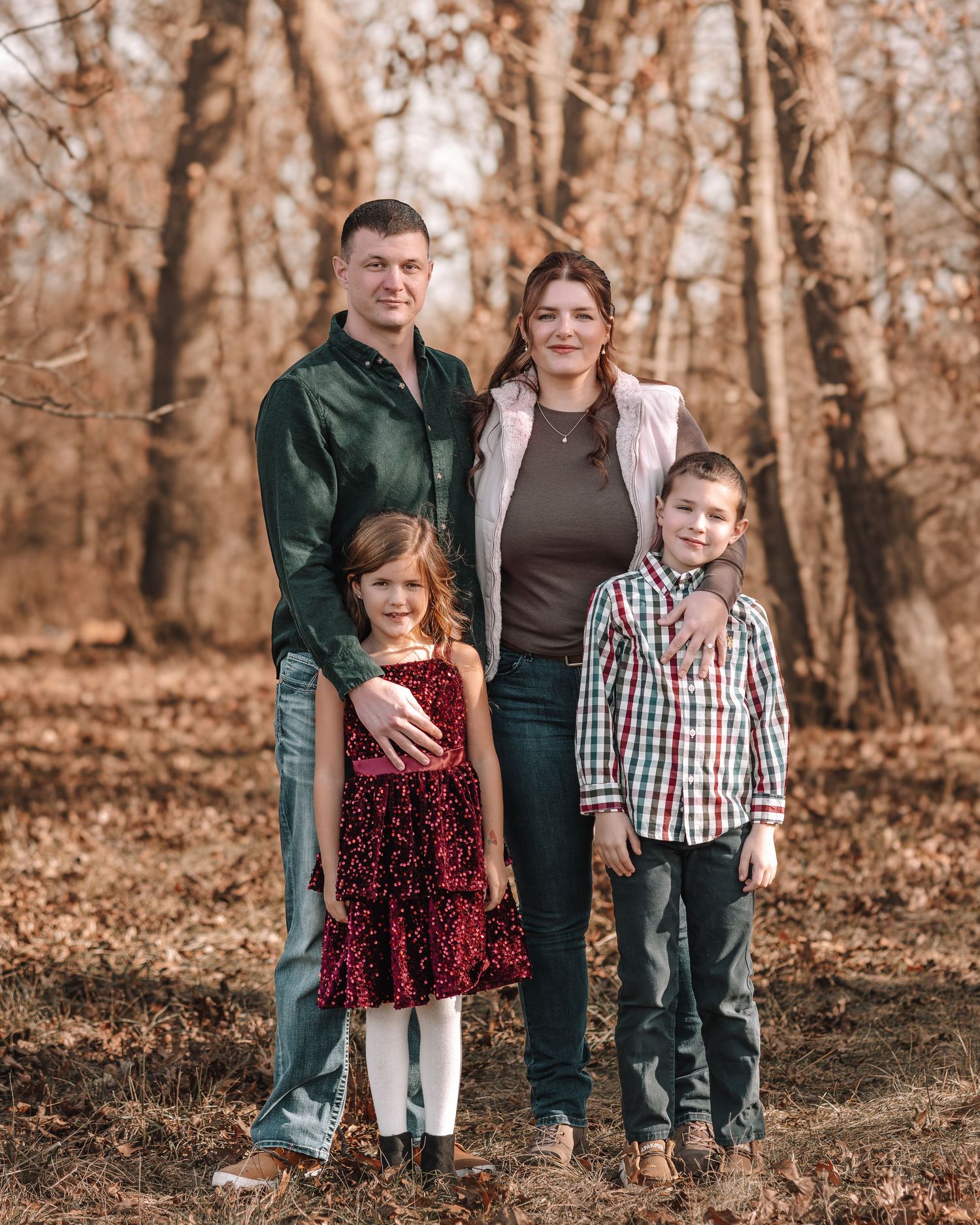 Family of four posing together for a portrait in a wooded area with fallen autumn leaves.