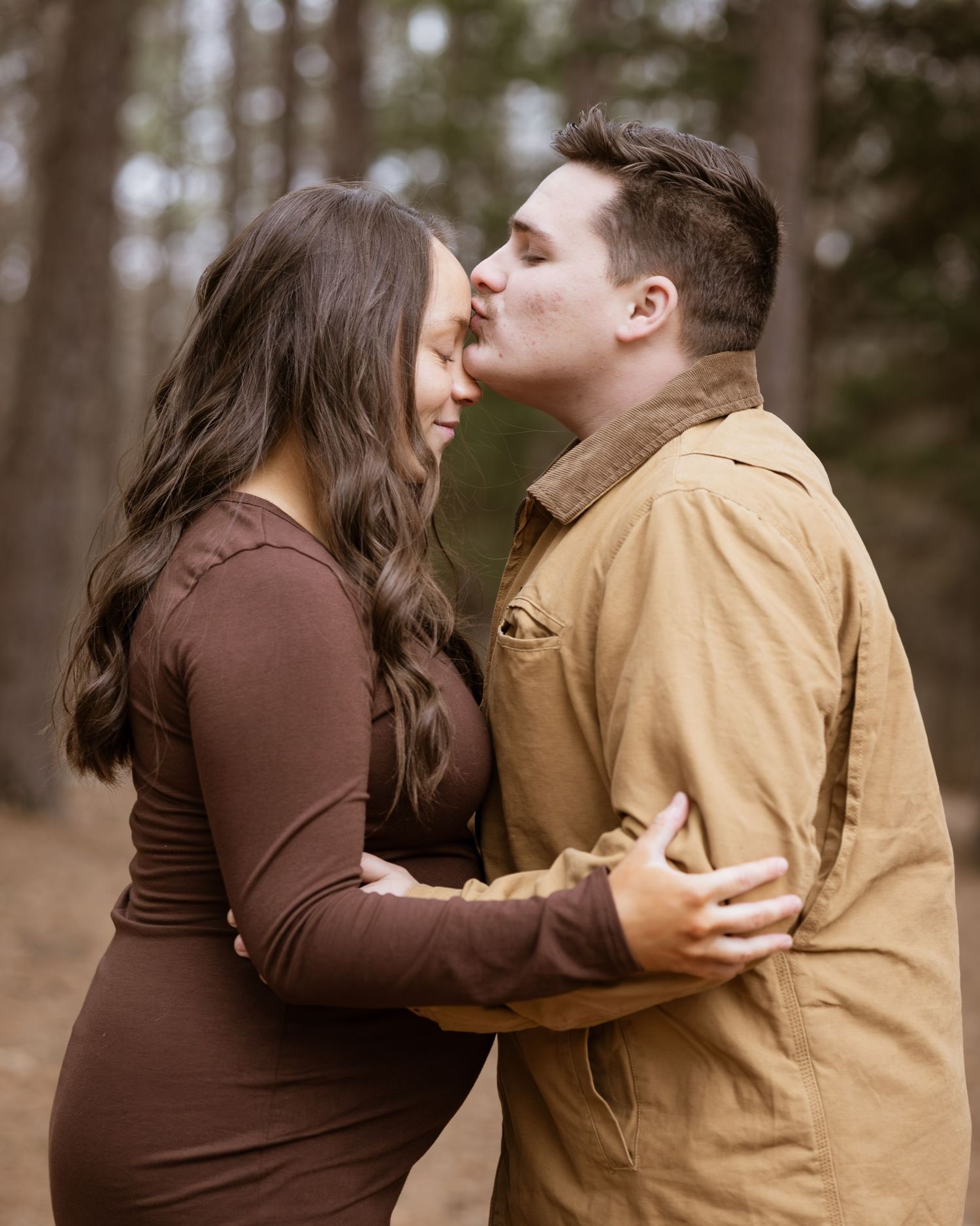 A romantic couple embraces in a forest setting wearing brown and tan clothing.