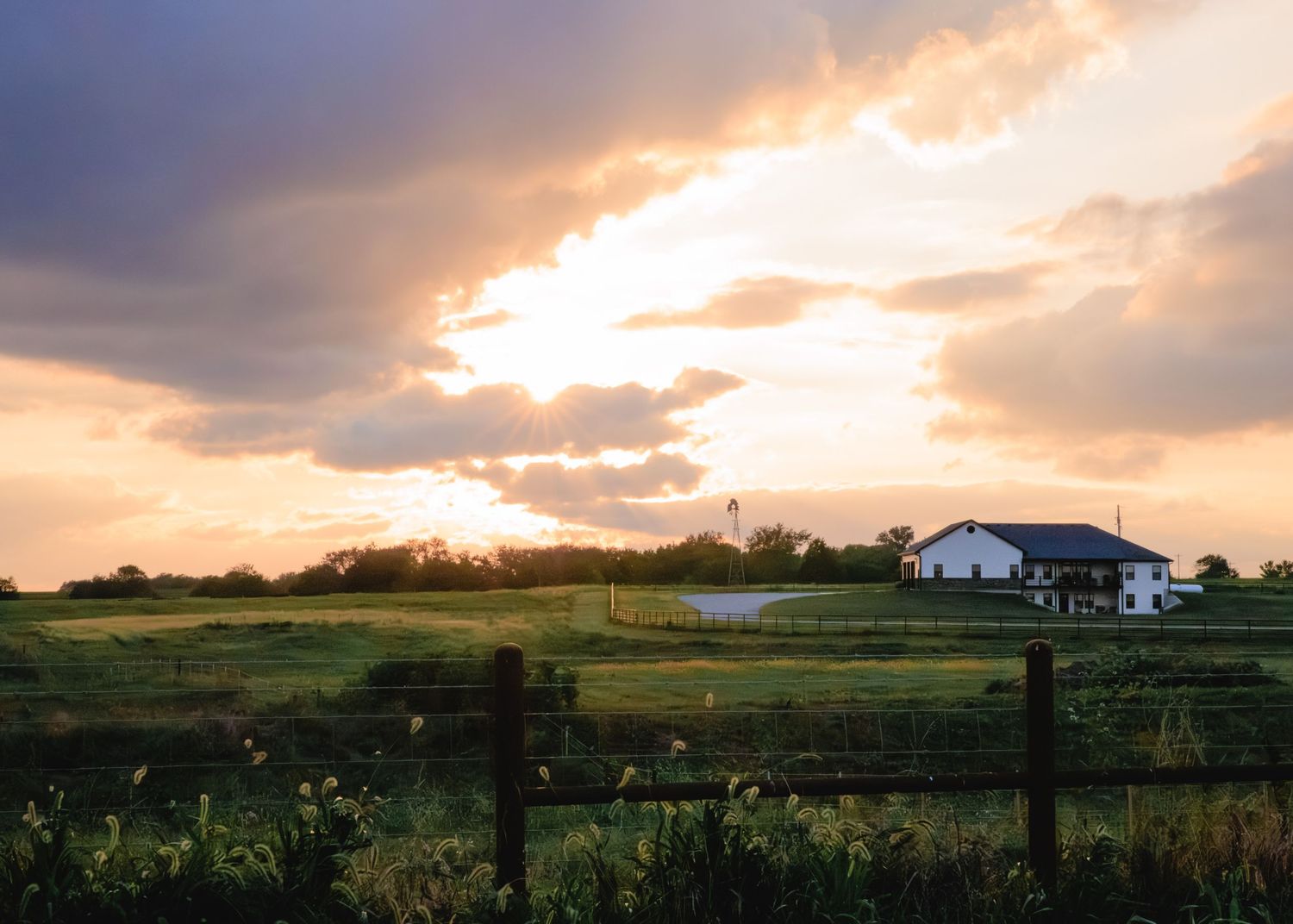Golden sunset over a rural farm with green fields, wooden fence, and white farmhouse under dramatic cloudy sky.