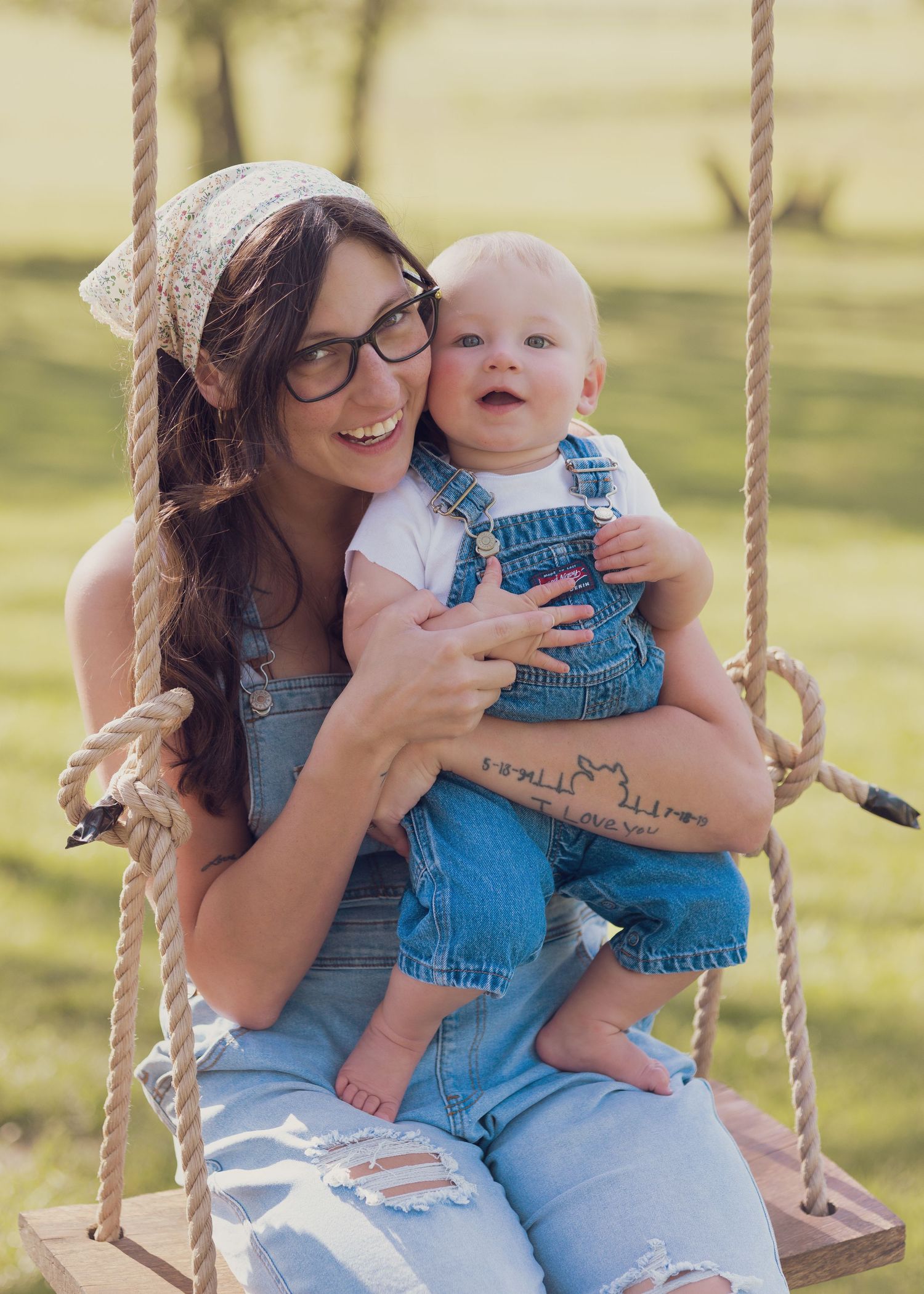 Young woman wearing glasses and overalls sits on a rope swing holding a smiling baby in denim overalls outdoors.