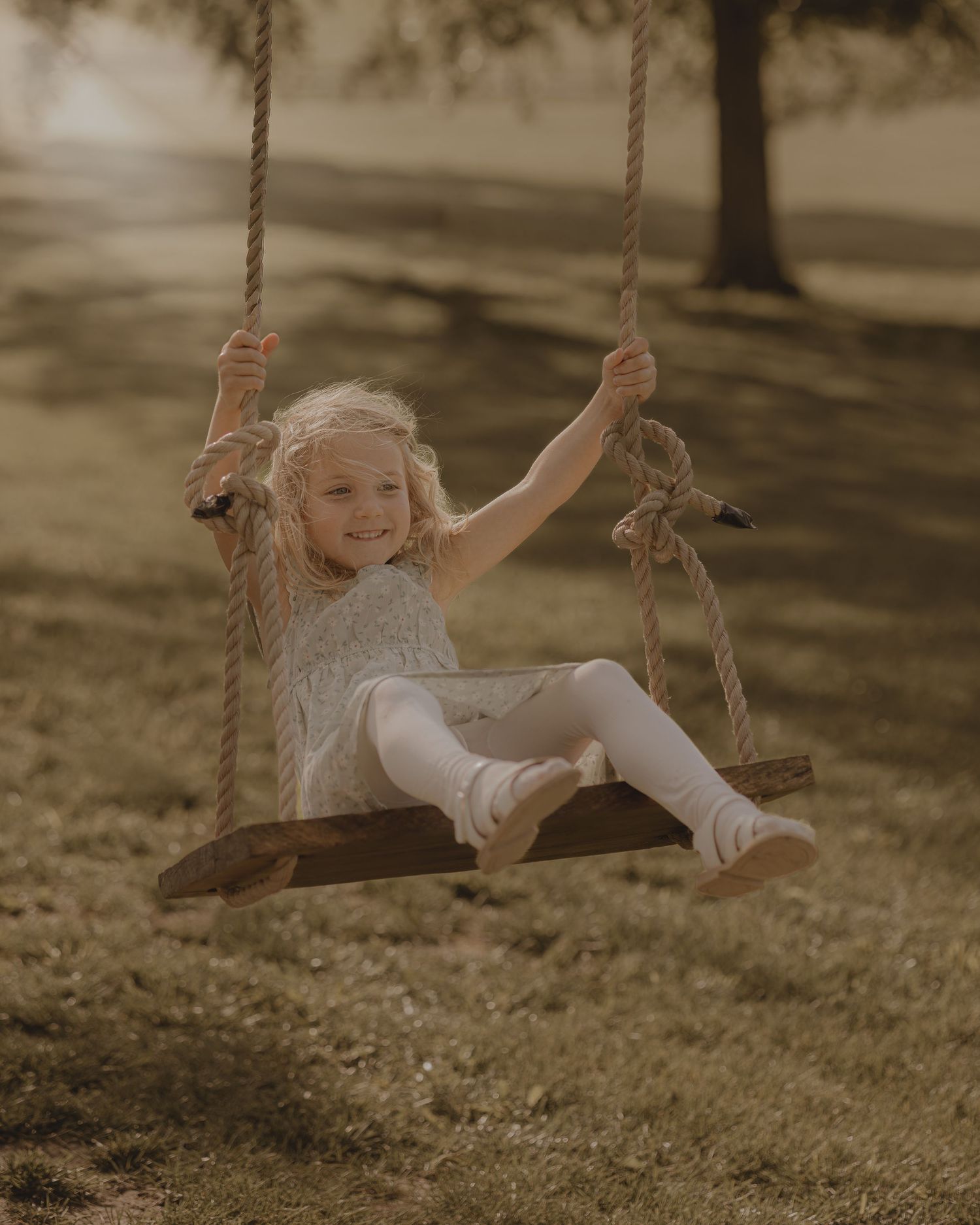 A happy blonde girl smiles while swinging on a rustic rope swing in a sunny outdoor park setting.