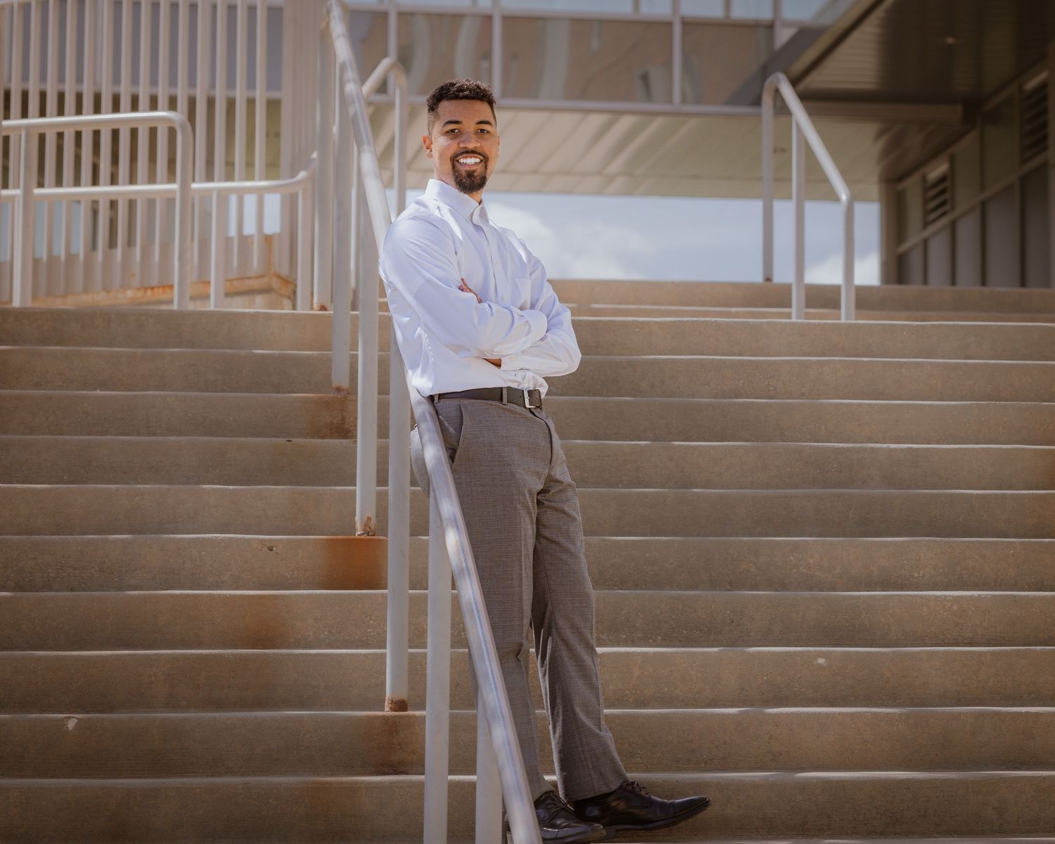 Professional man in white shirt and gray trousers standing confidently on outdoor stairs with arms crossed.