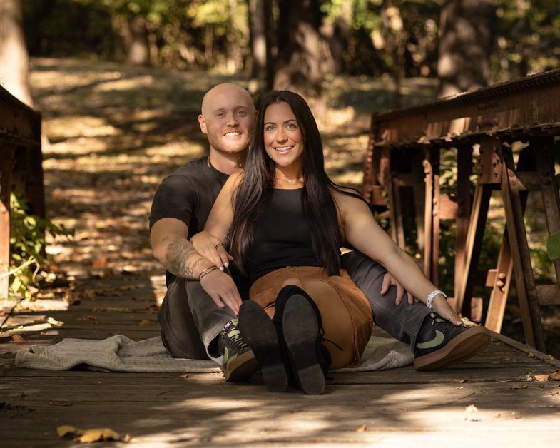 Couple sitting together on a wooden bridge in a sunlit forest, smiling and posing for an outdoor portrait photo.