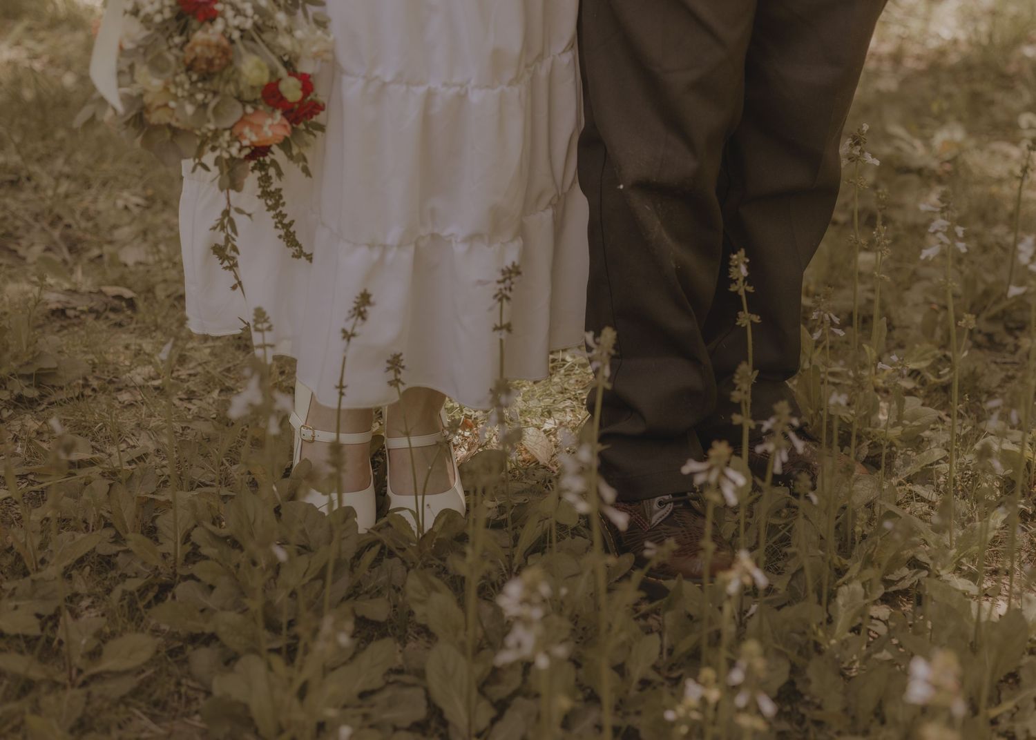 Bride in white dress and groom in dark suit standing in wildflower field, wedding couple close-up.