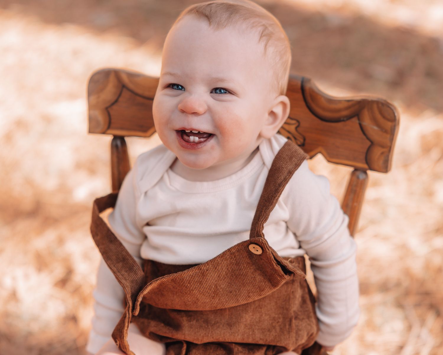 A small child sits in a wooden chair wearing brown overalls in a forest setting.
