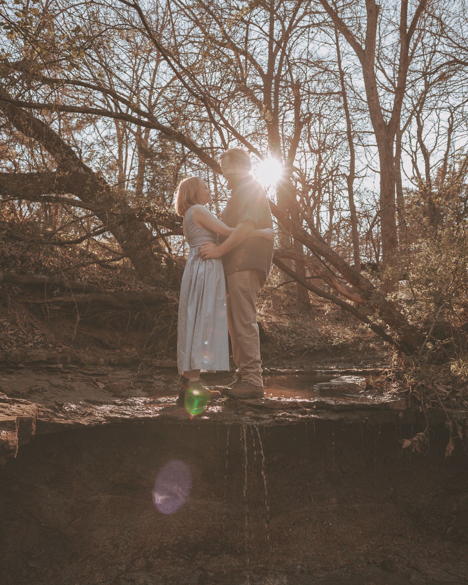 Couple embracing at sunset in a forest, sun flare through bare trees, romantic outdoor engagement photo.