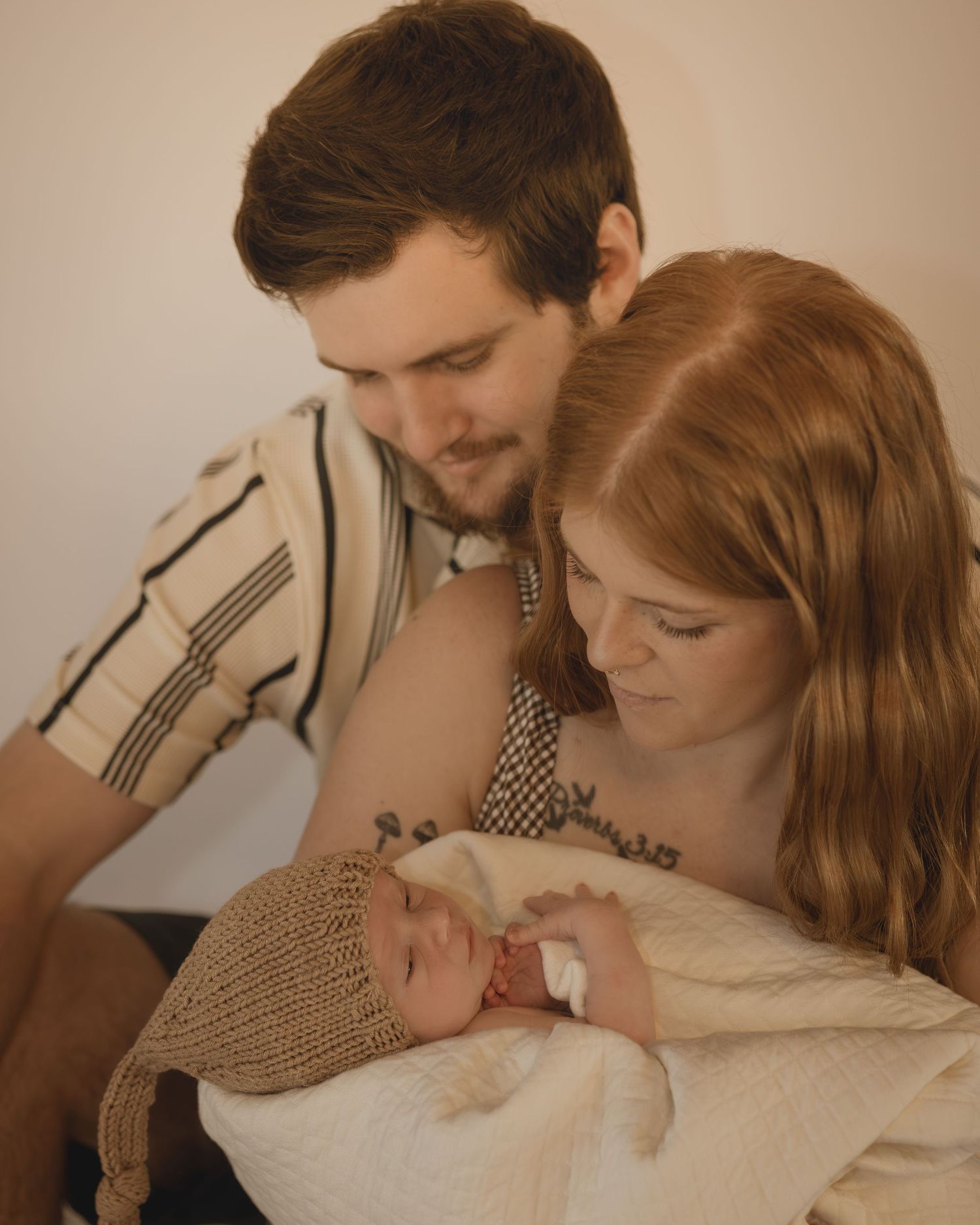 Young couple gazing lovingly at their newborn baby wrapped in cream blanket with knit hat.