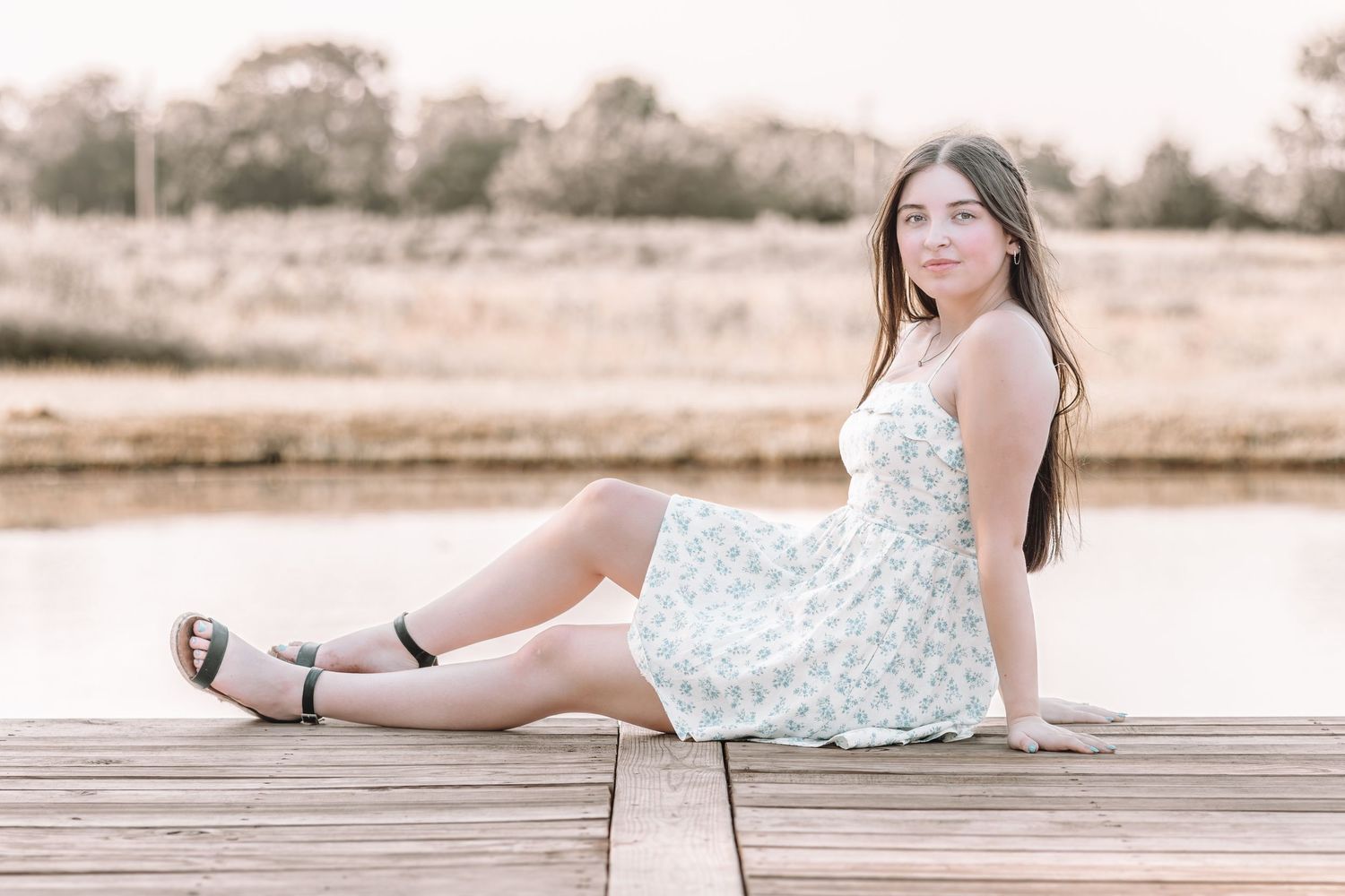 A figure in a white floral dress poses gracefully on a wooden dock by a serene lake at sunset.
