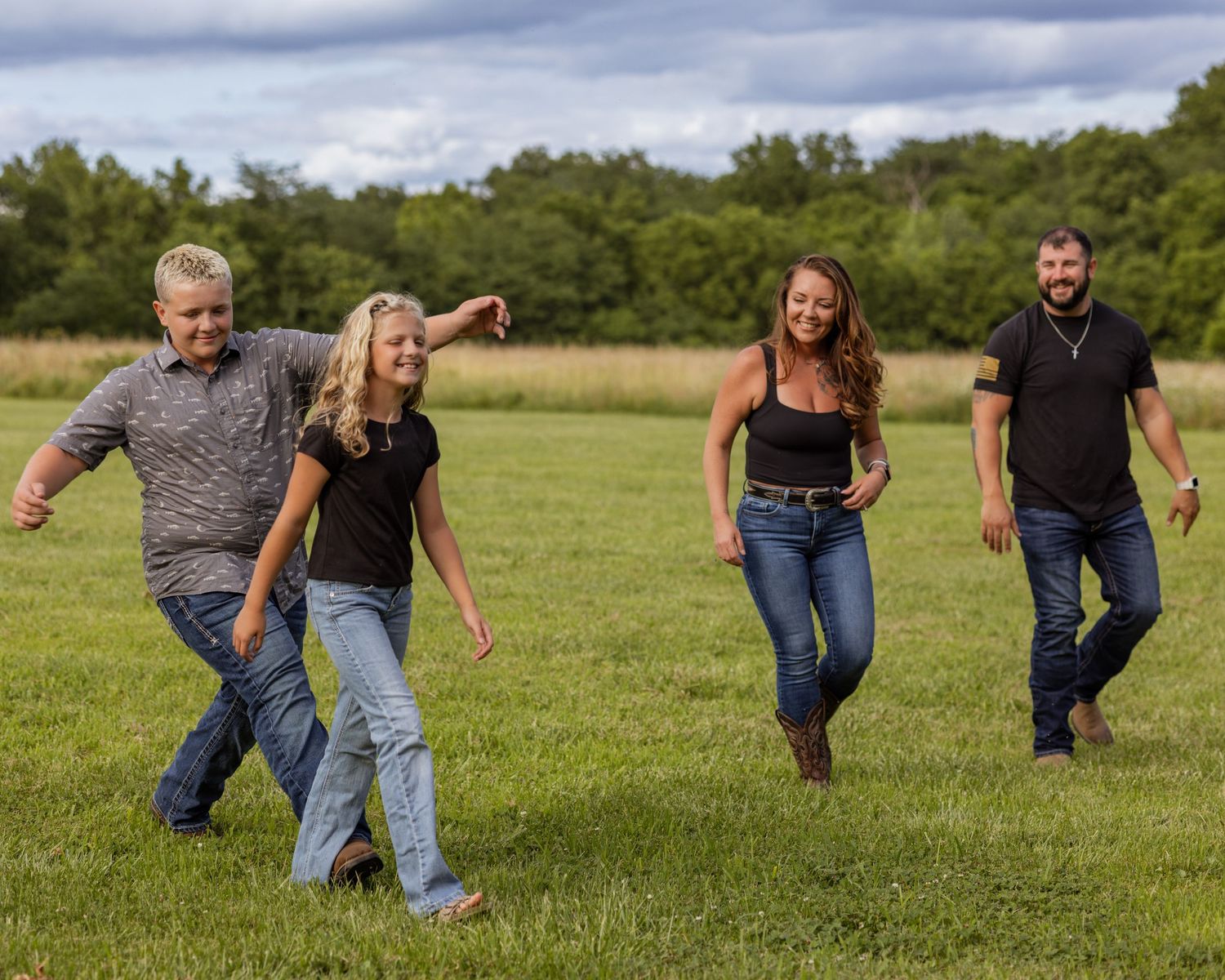 Two couples walk and laugh together in a lush green field under a cloudy sky.