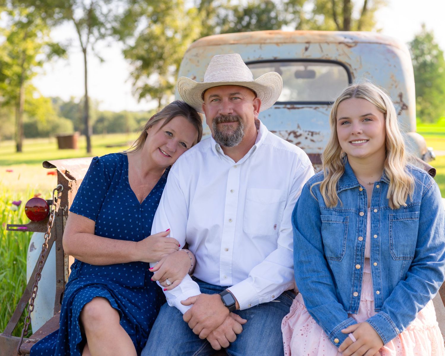 Family of three smiling together outdoors by a vintage truck on a sunny farm day.