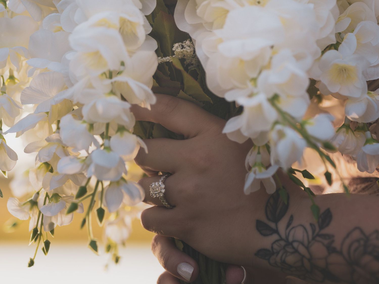 Tattooed hands holding white orchid wedding bouquet with diamond engagement ring, close-up bridal detail shot.