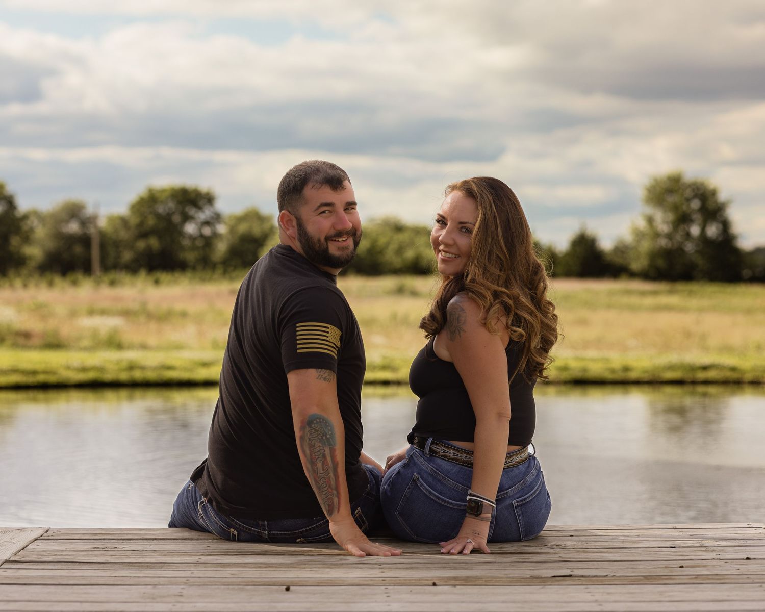 Couple sitting back-to-back on a wooden dock by a pond, smiling, man with tattoos in black tee, woman in black top and jeans.