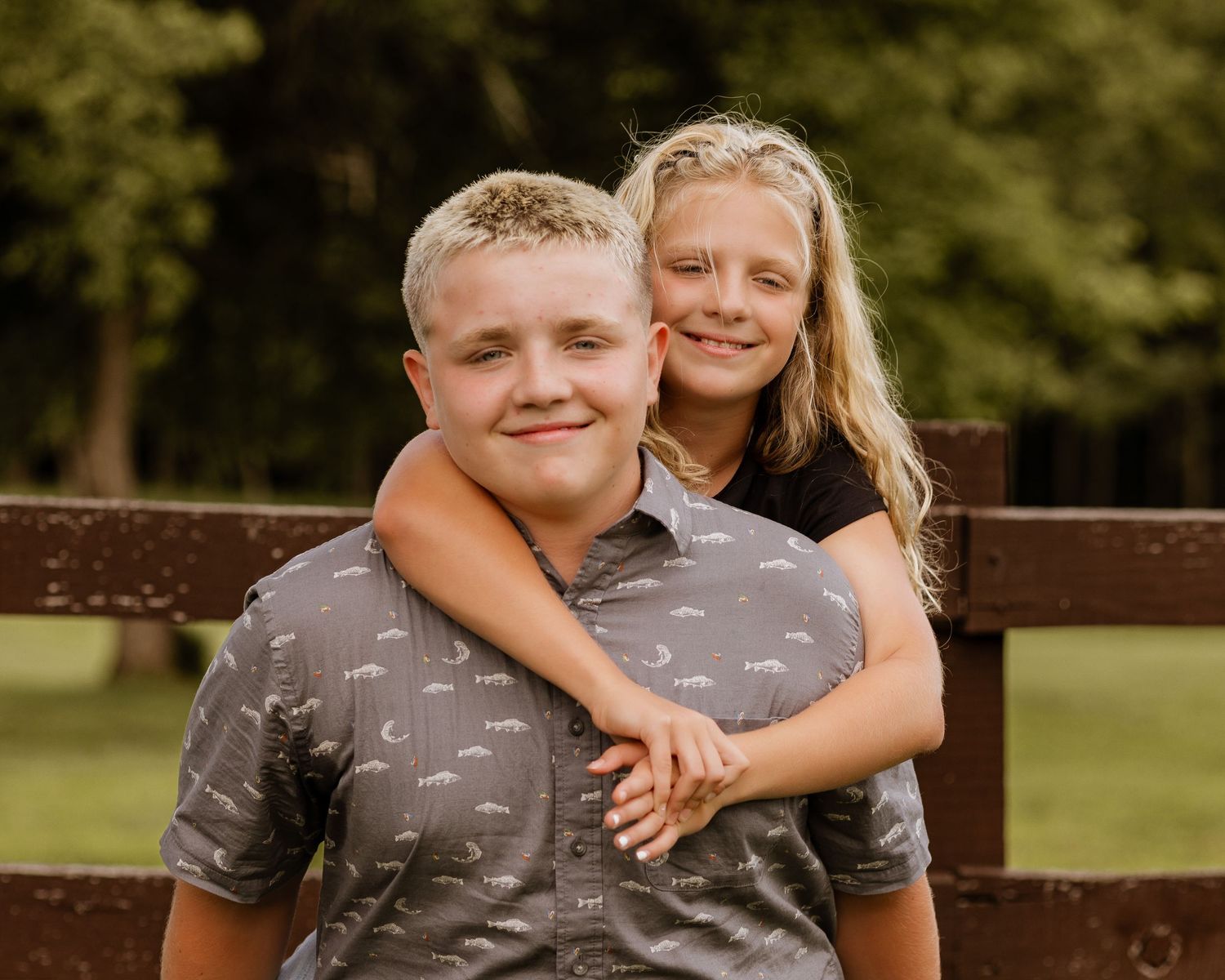 Two smiling siblings, a boy and girl, posing together outdoors near a wooden fence in a park.
