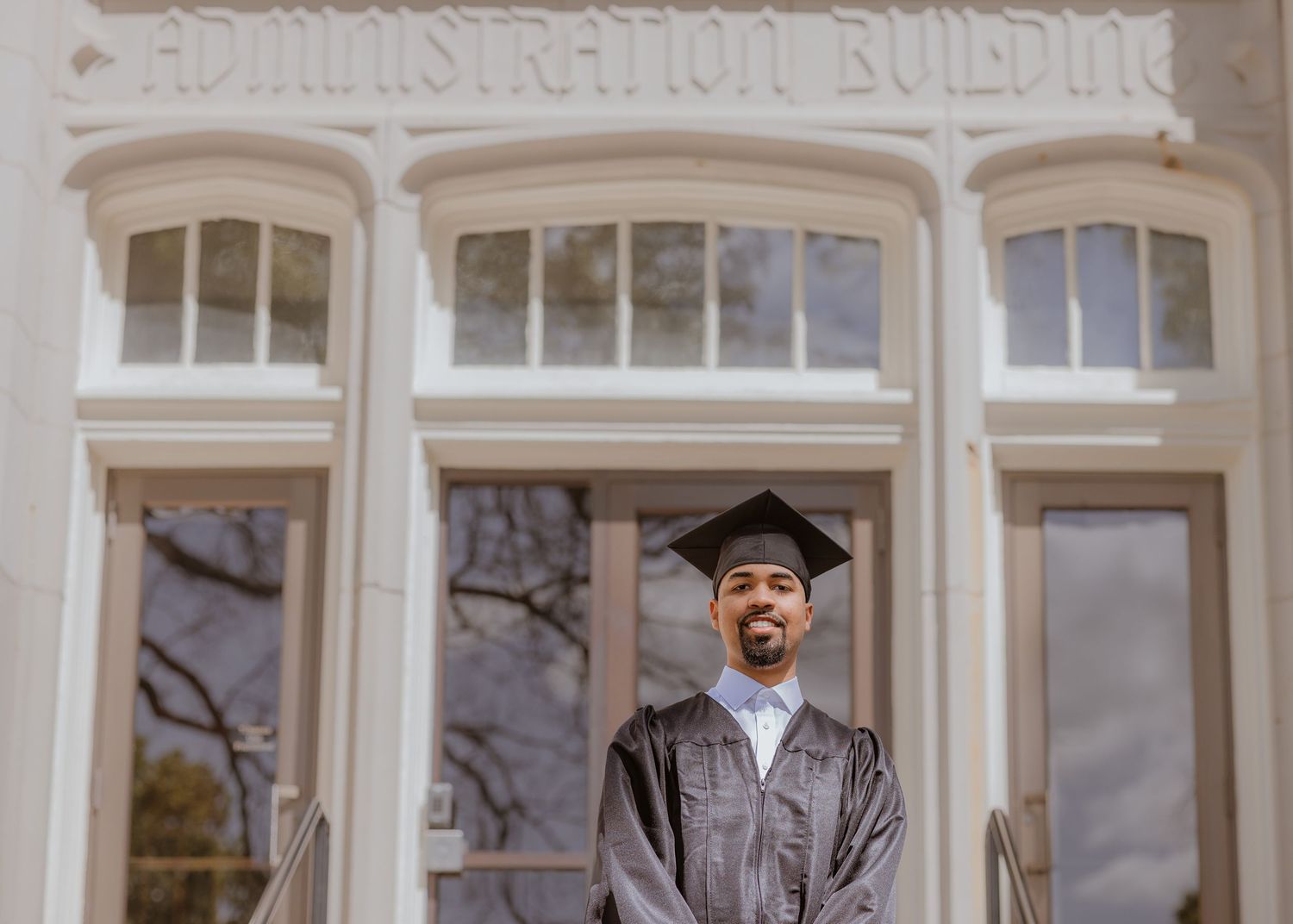 Graduate in cap and gown smiling in front of Administration Building on college campus graduation day.