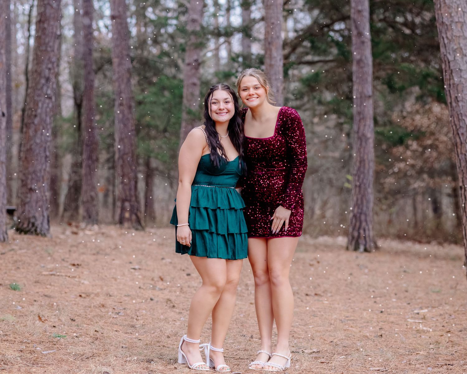 Two people in formal dresses pose together in a forest setting with tall pine trees in the background.