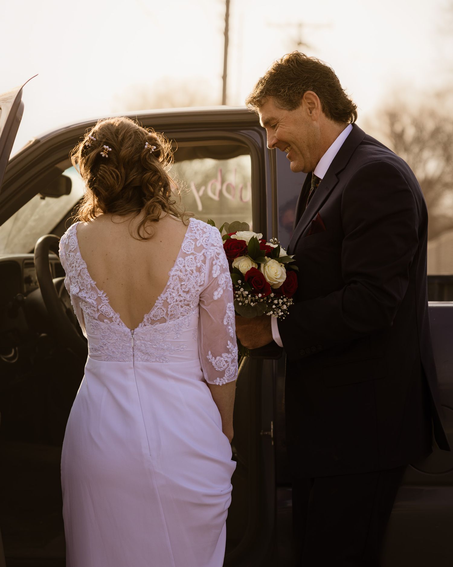 Bride in white lace dress stands next to car with elegant details on a sunny day.