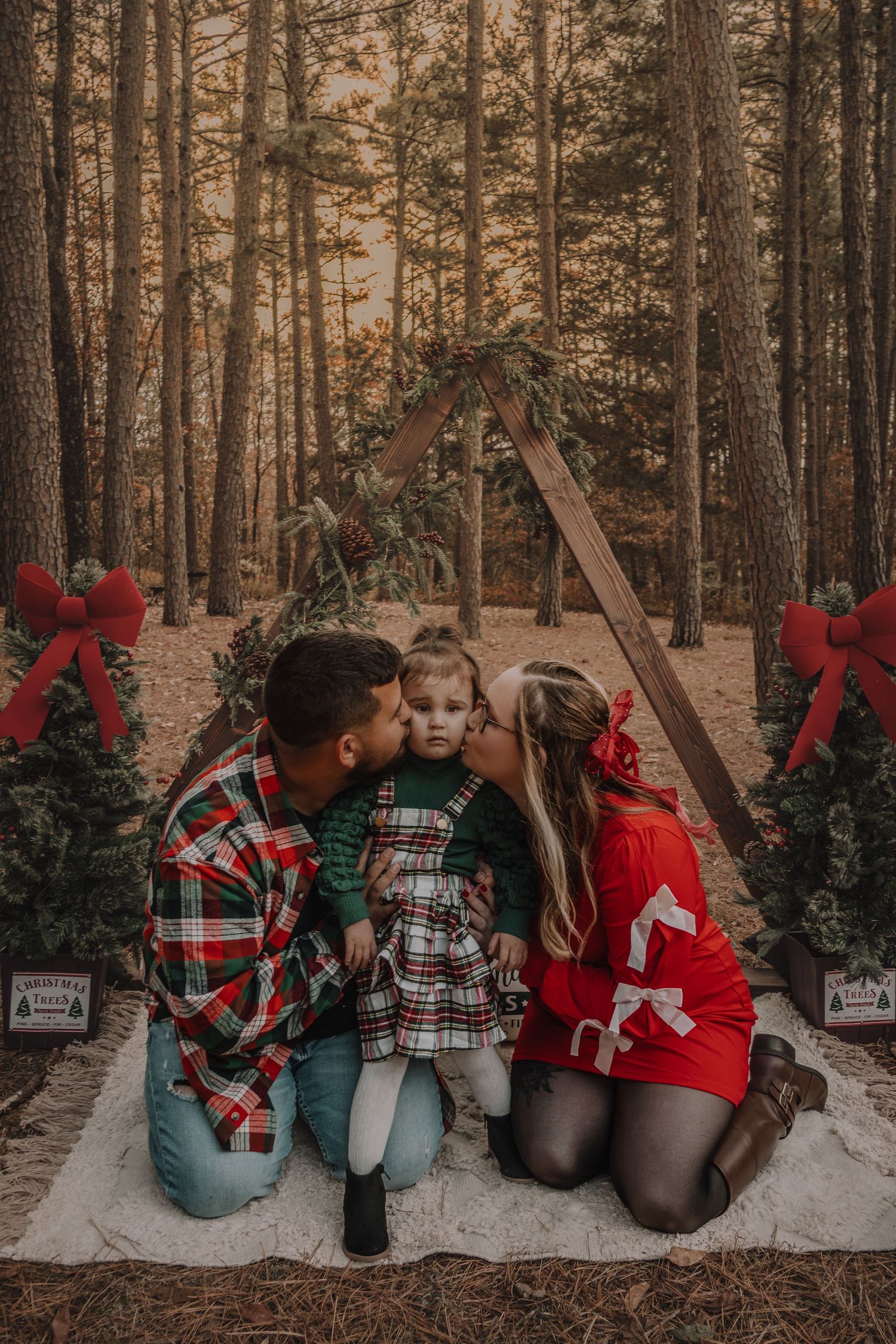 Family in matching holiday outfits sitting in snowy forest with Christmas trees and decorative red bows.