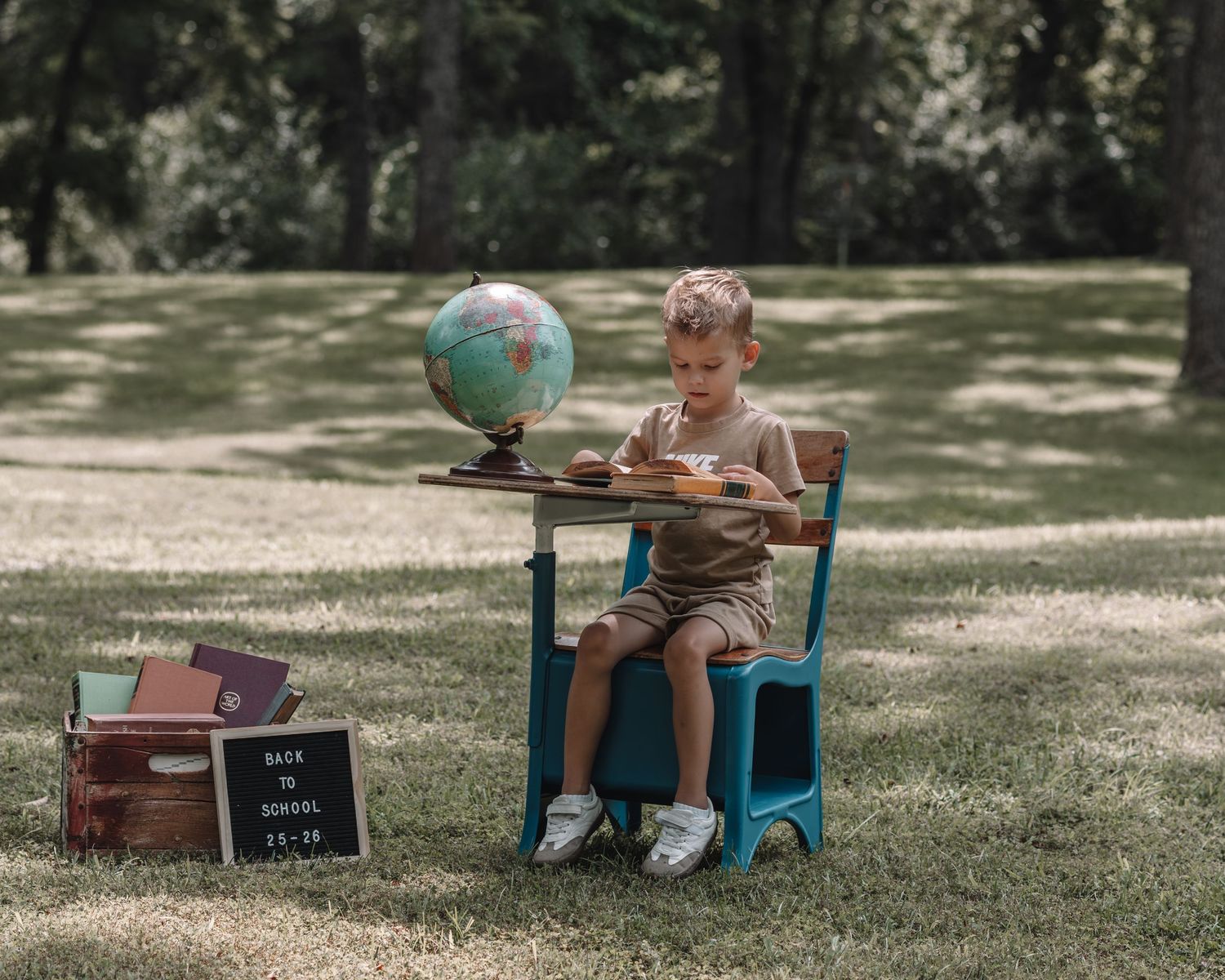 A child sits in a blue chair on grass with vintage suitcases and a globe, creating a travel-themed photo setup.
