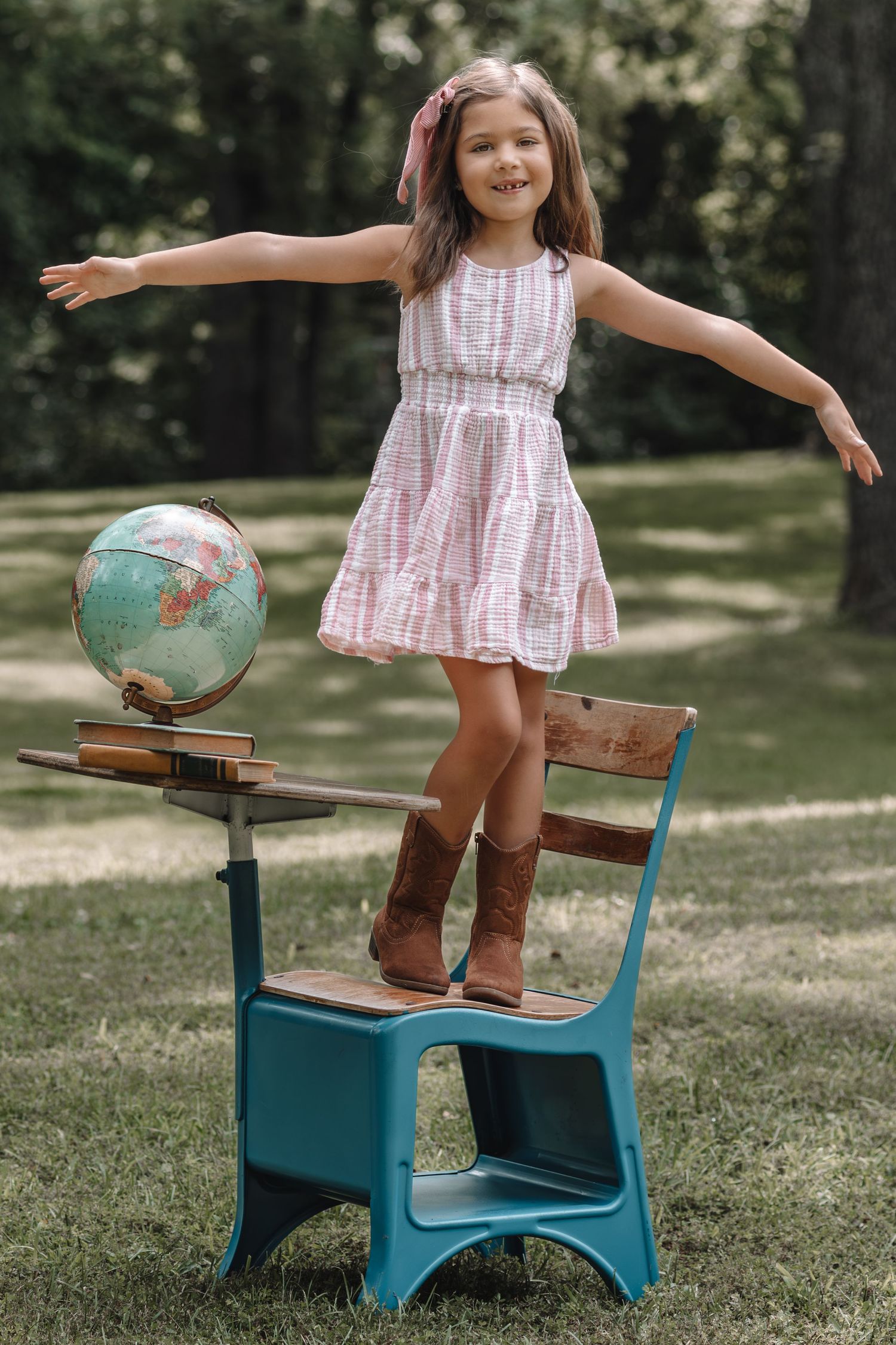 A child in a pink dress and brown boots stands playfully on a vintage turquoise school desk with a globe in an outdoor setting.