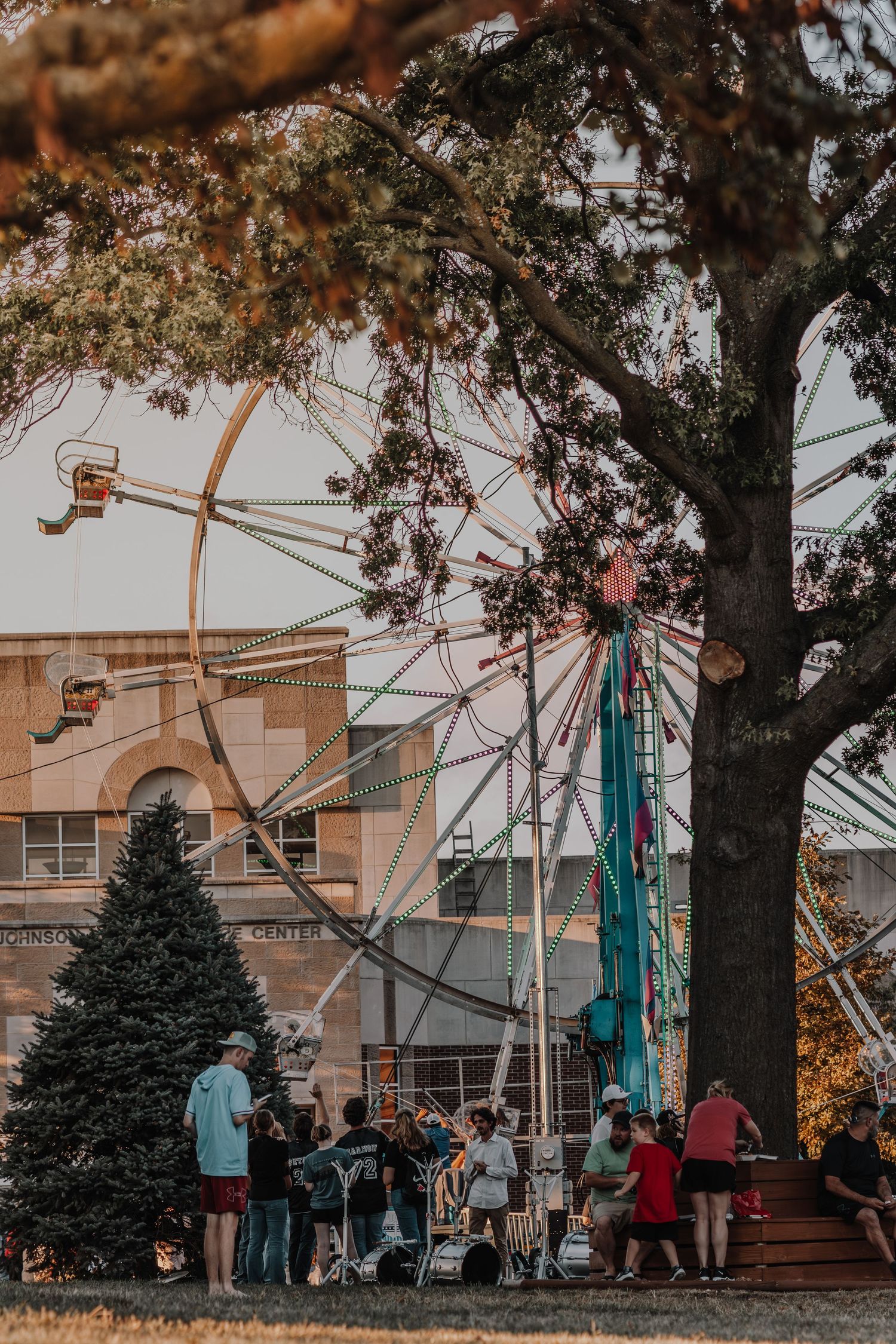 People gather under autumn trees near a building with ski lift chairs visible in an outdoor social setting.