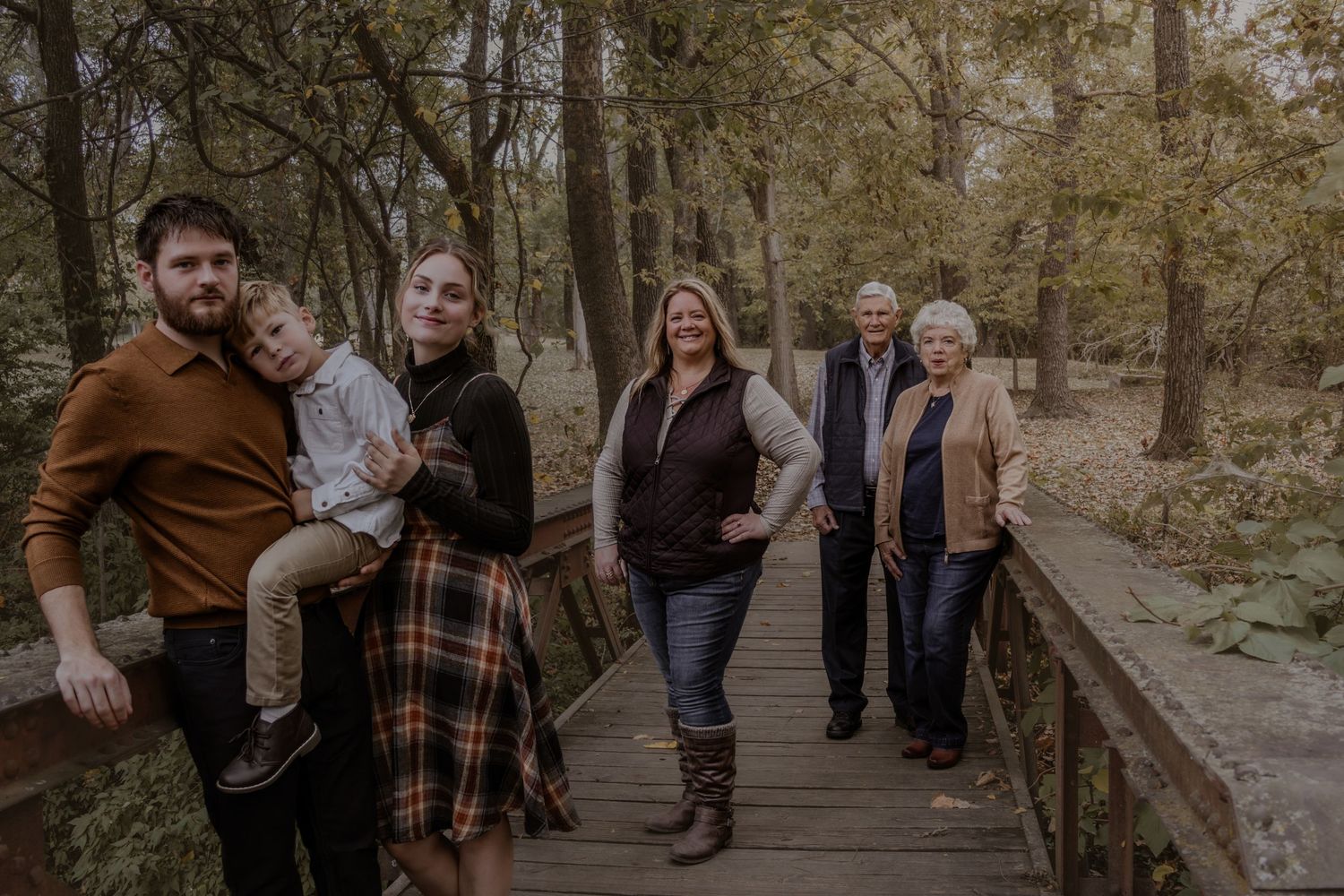 Multi-generational family in fall attire poses together on a wooden bridge in an autumn forest setting.