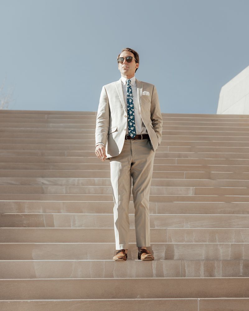A person in a beige suit and sunglasses stands on wide concrete steps against a bright sky.