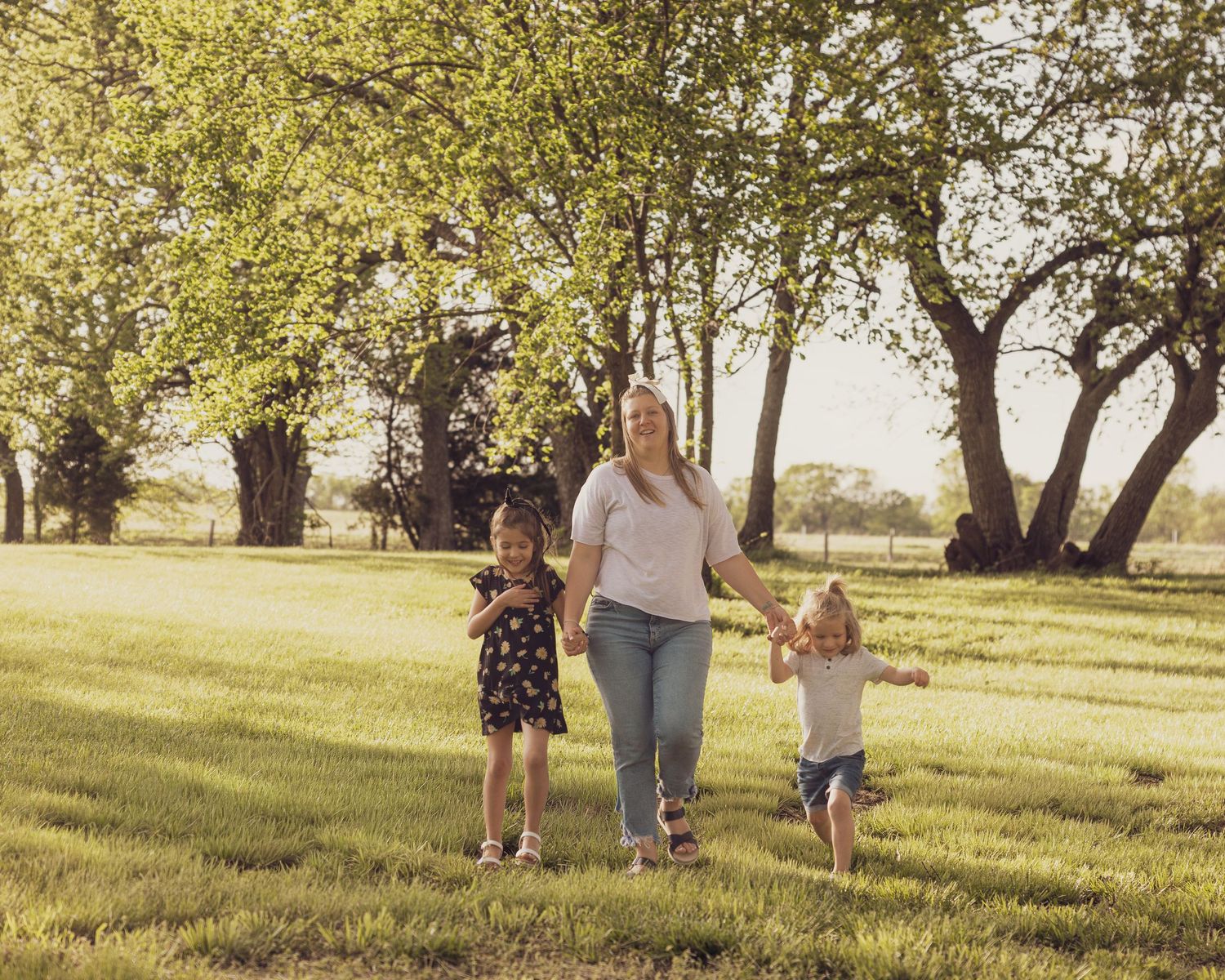 A woman walks hand-in-hand with two young girls across a sunny green park surrounded by large trees.