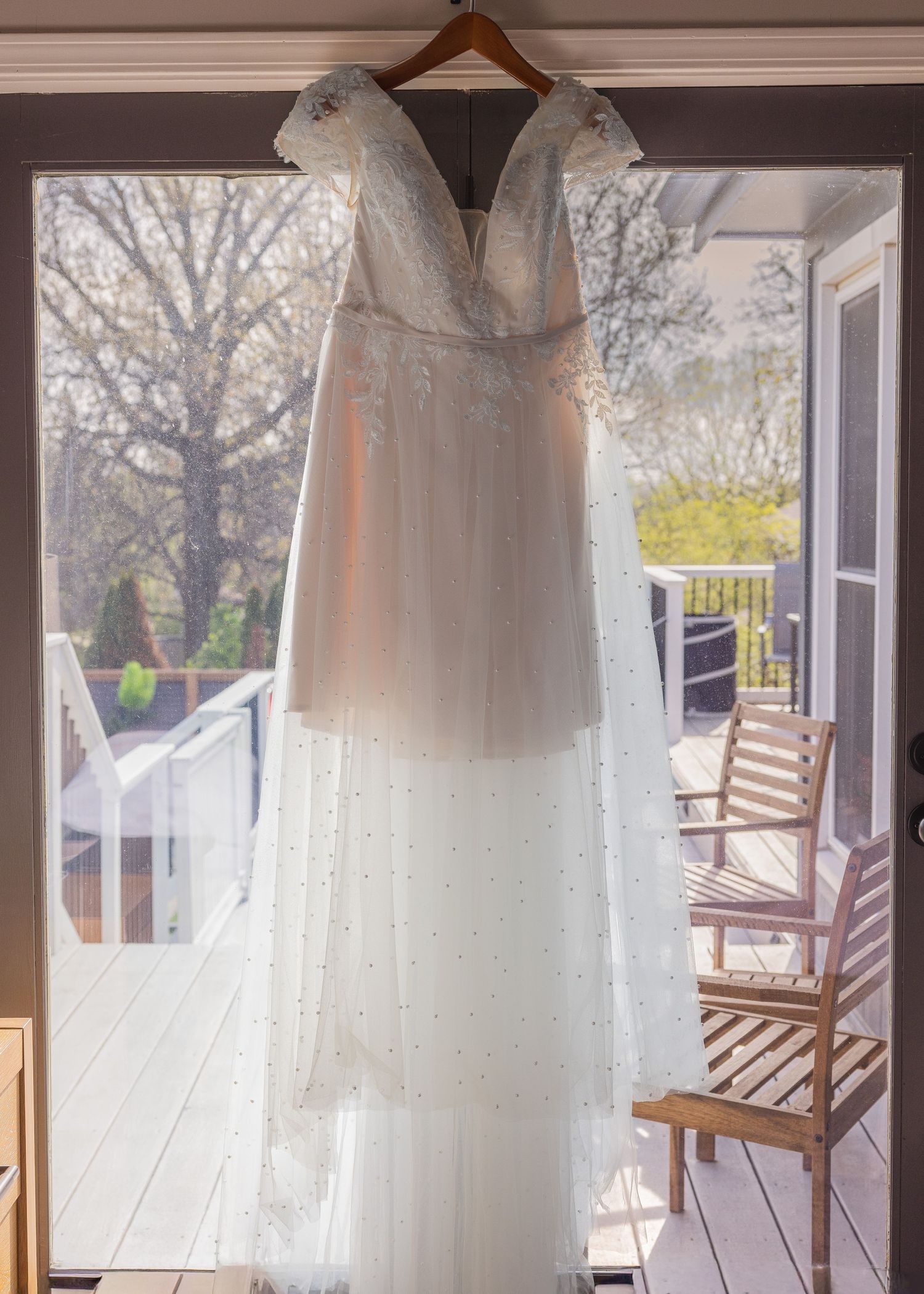 Elegant white beaded wedding gown with cap sleeves hanging in a sunlit window overlooking a wooden deck.