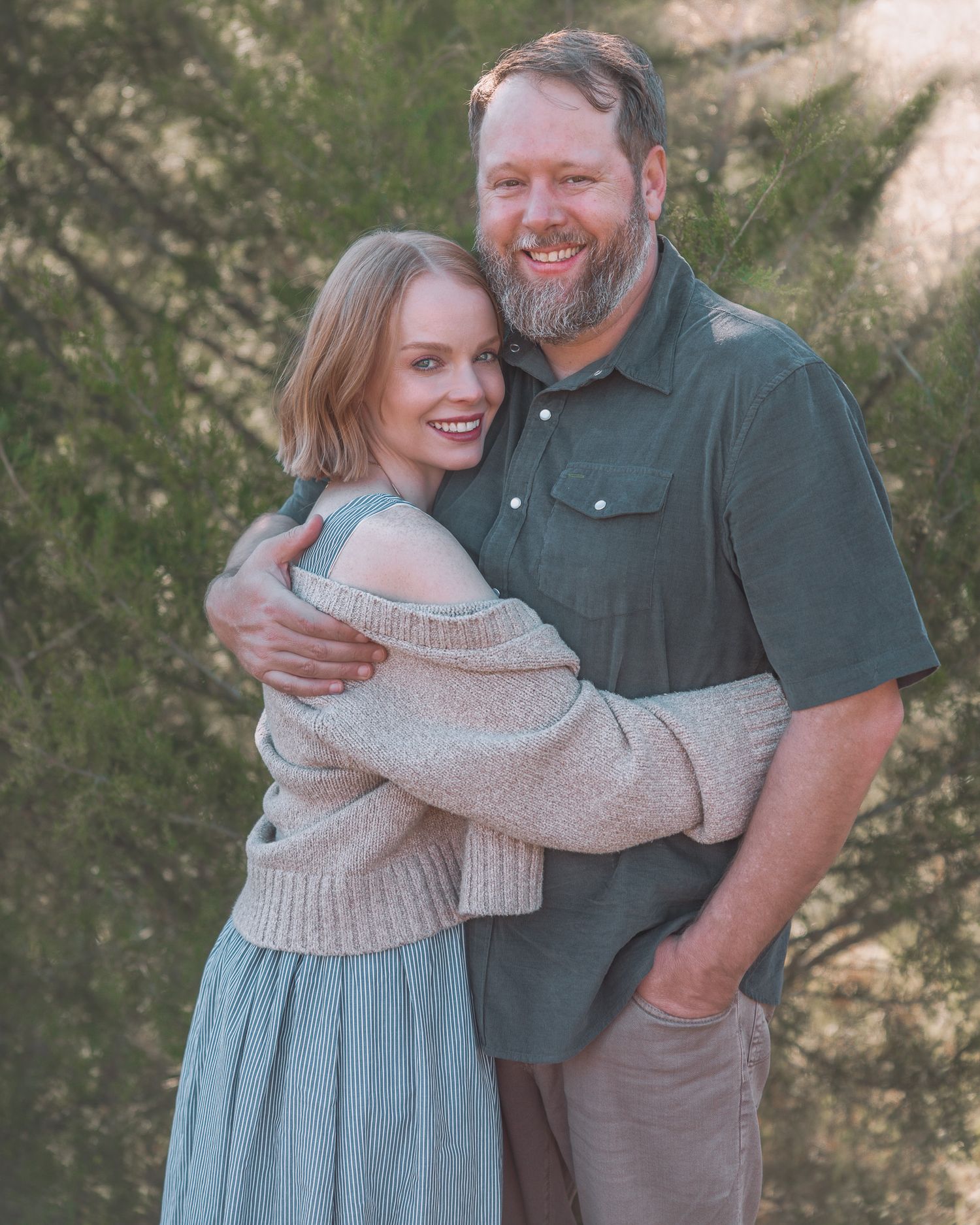 Smiling couple embracing warmly outdoors in front of green evergreen trees.