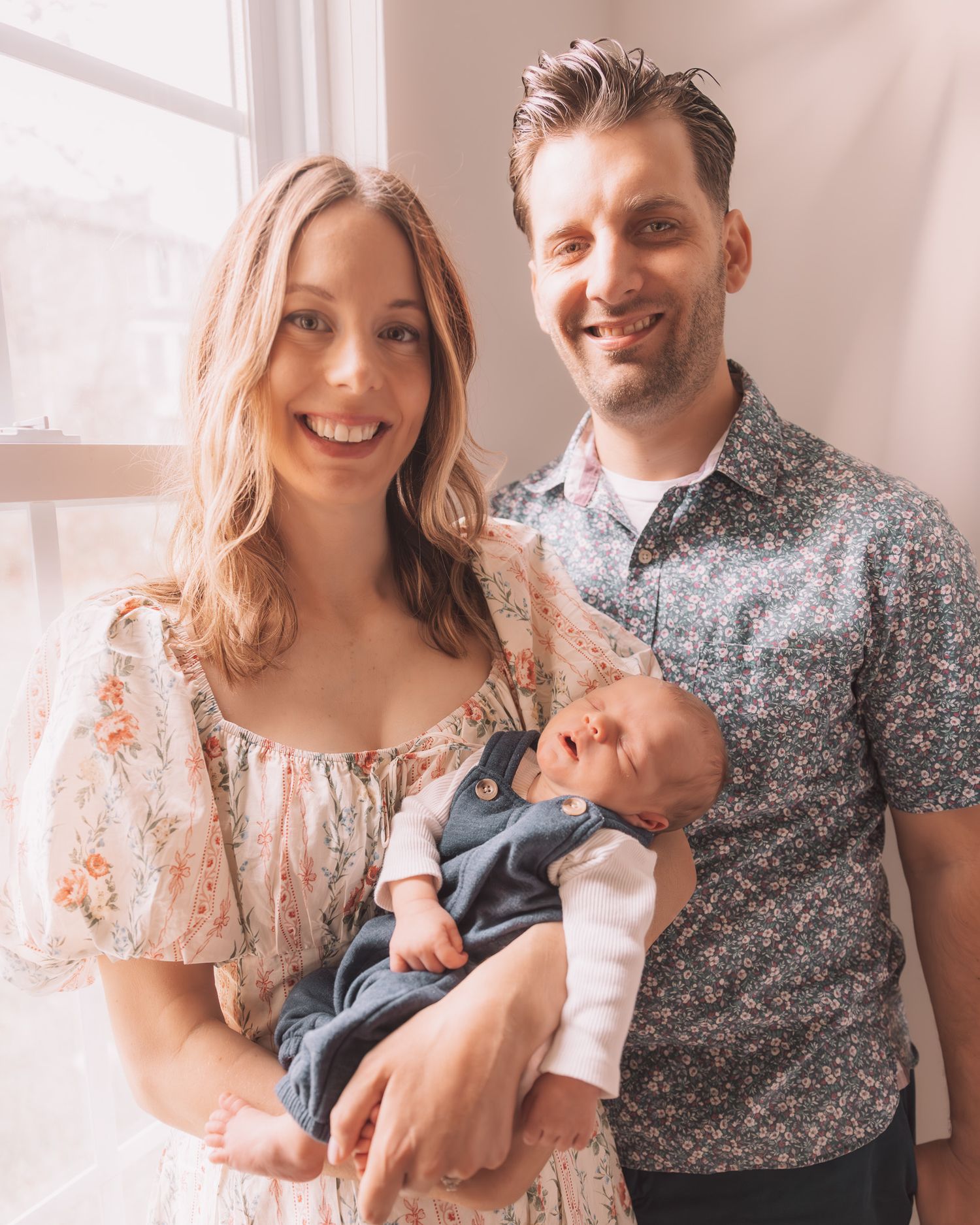 Smiling couple posing with their newborn baby by a bright window in floral outfits.