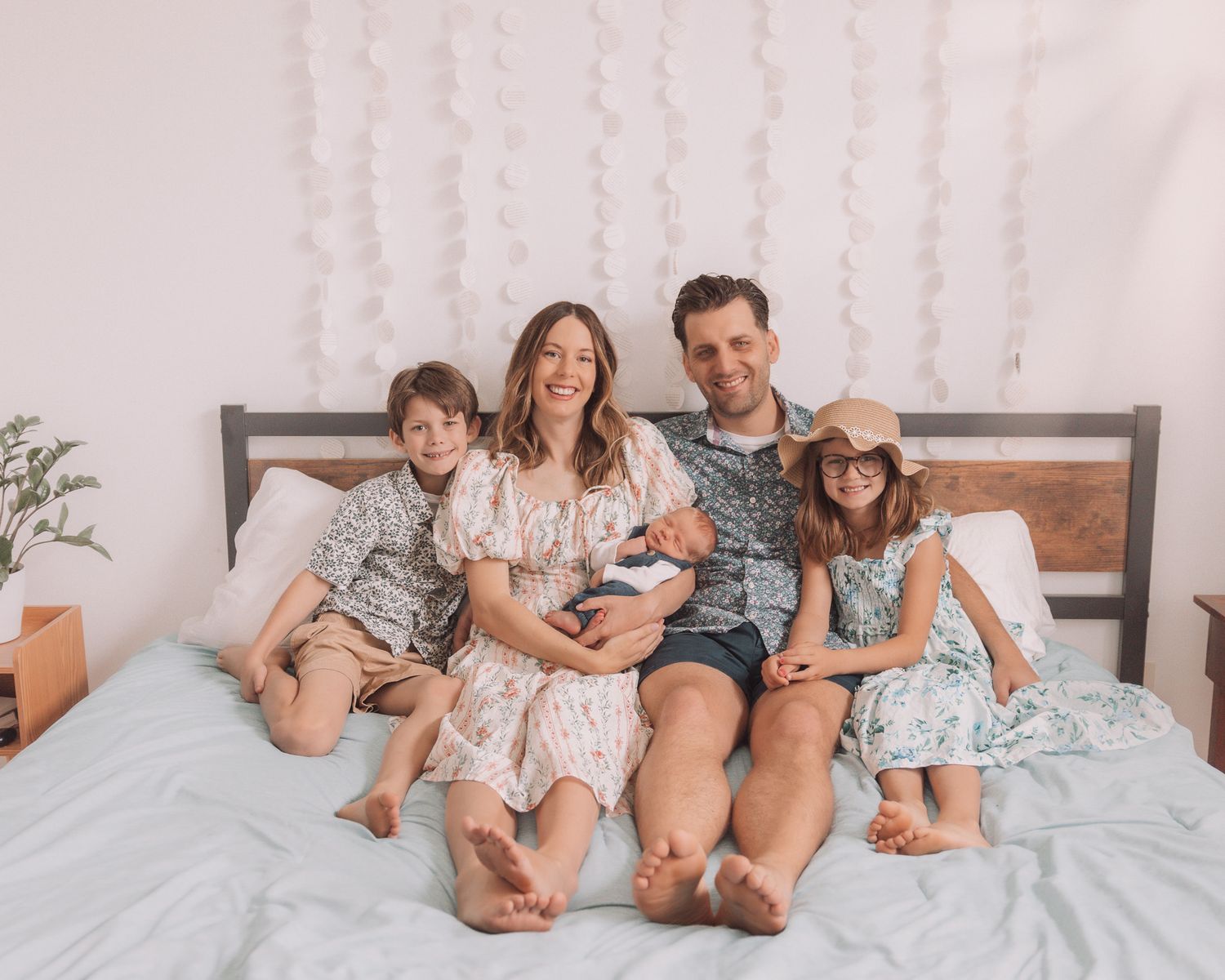 A family of five sitting on a bed, parents holding a newborn with two older children beside them.