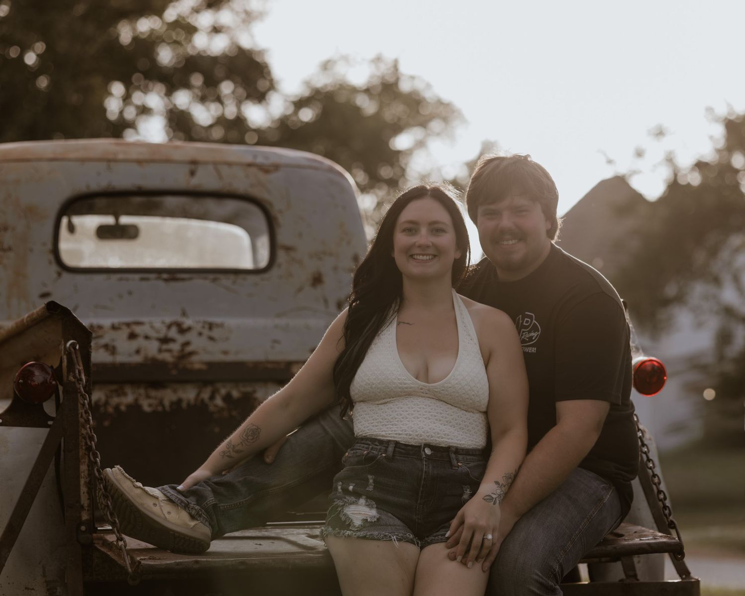 A smiling couple poses on the back of a vintage rusty truck during a warm golden hour photo session.