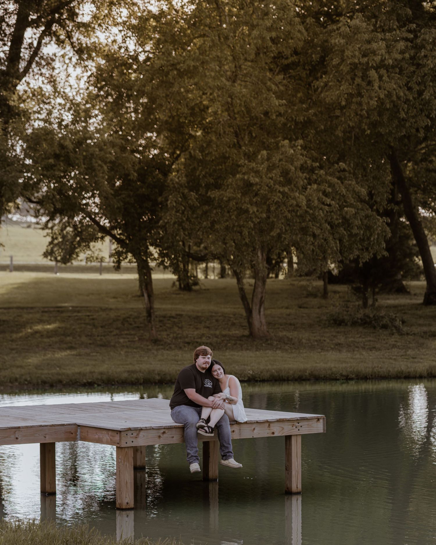 Couple sitting together on a wooden dock by a calm pond surrounded by lush green trees.