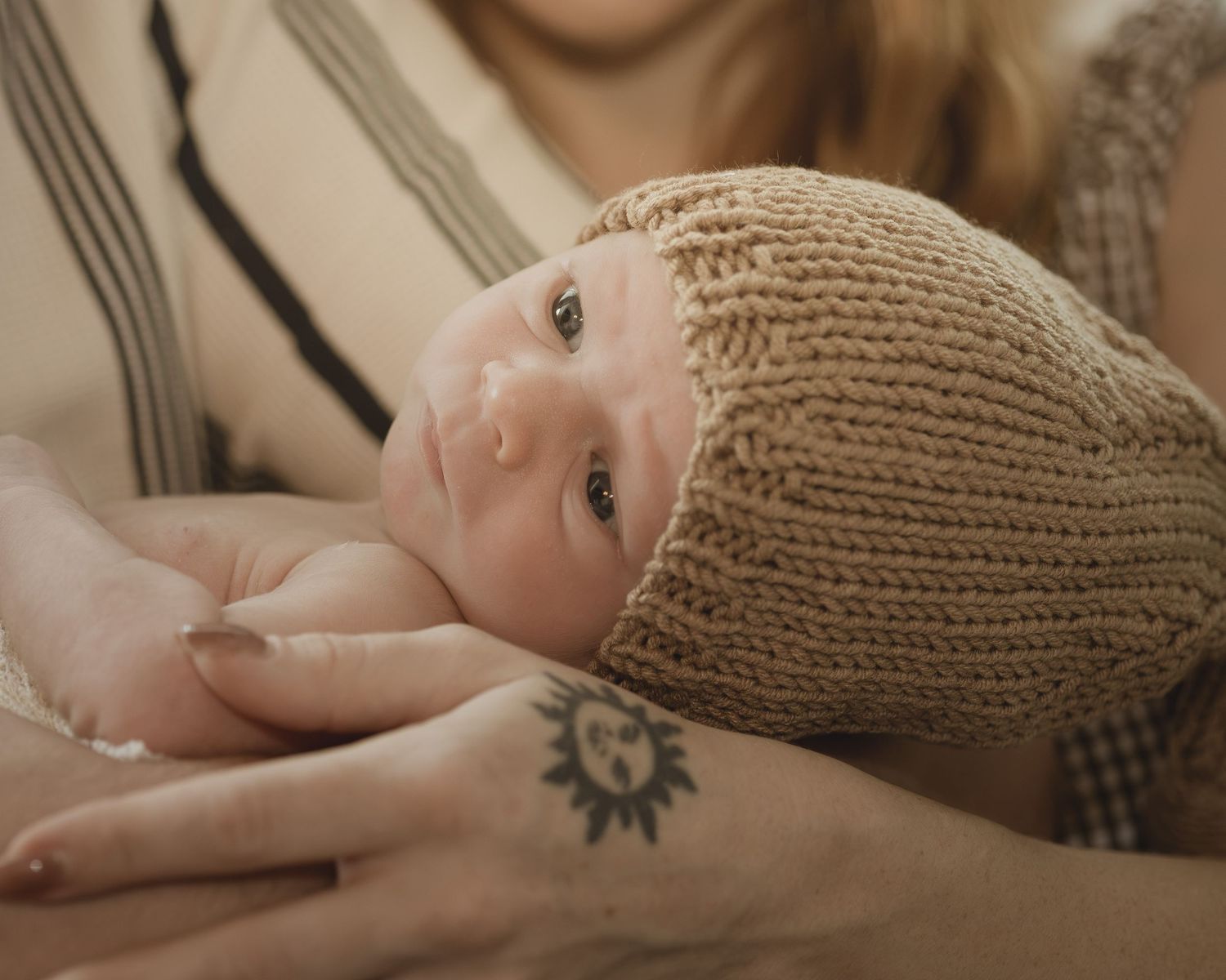 Newborn baby in knit beige hat cradled in parent's arms, with sun tattoo visible on wrist.