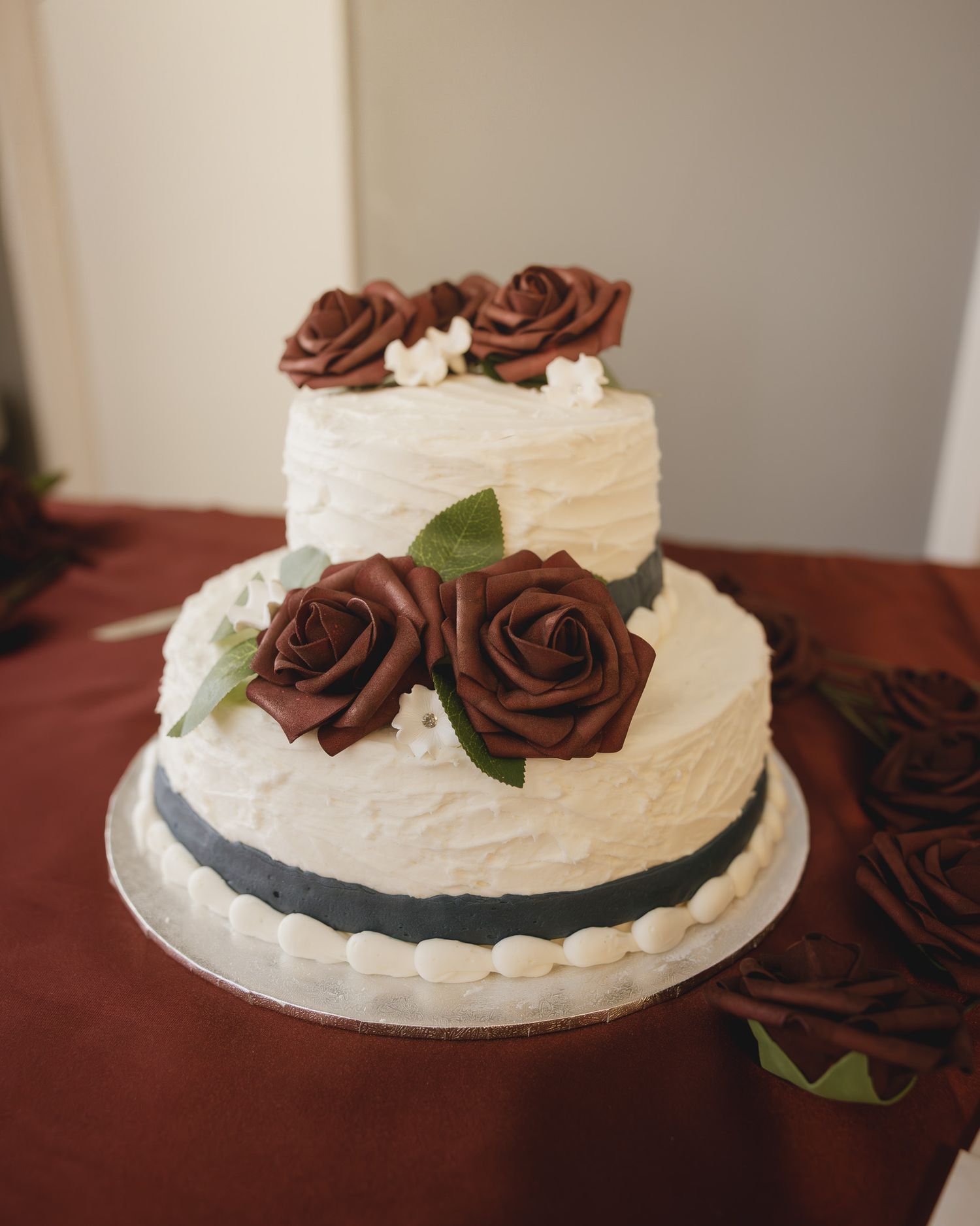 Two-tier white wedding cake with chocolate rose decorations and gray ribbon accents on a silver cake board.