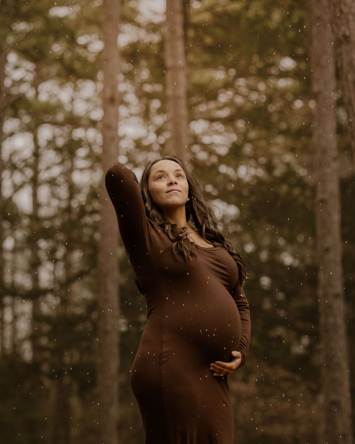 Pregnant woman in brown dress holding belly among forest trees with raindrops falling around her.