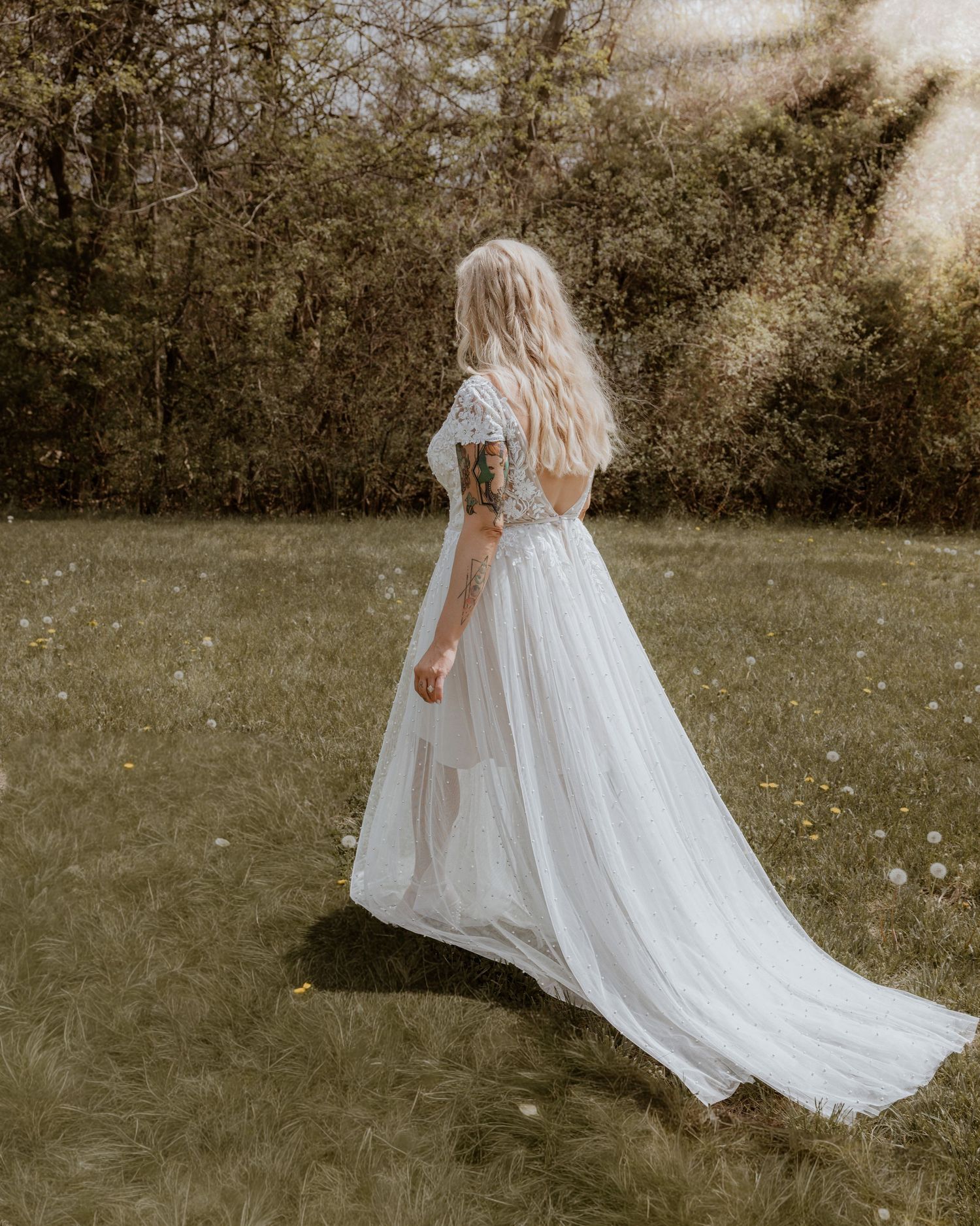 Bride in flowing white wedding gown with lace top walks through green meadow with train trailing.