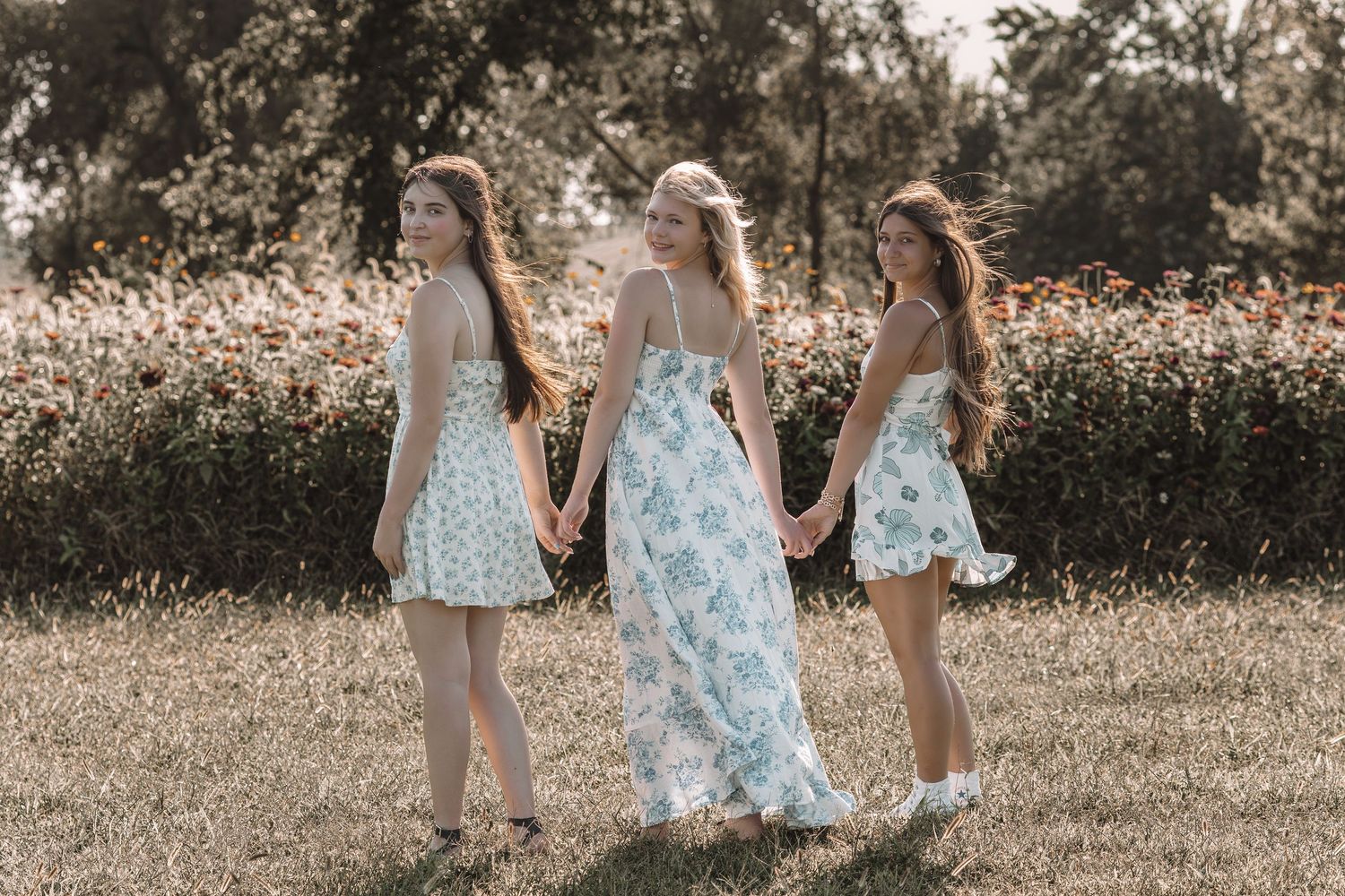 Three people in floral dresses walking together through a sunny outdoor field.