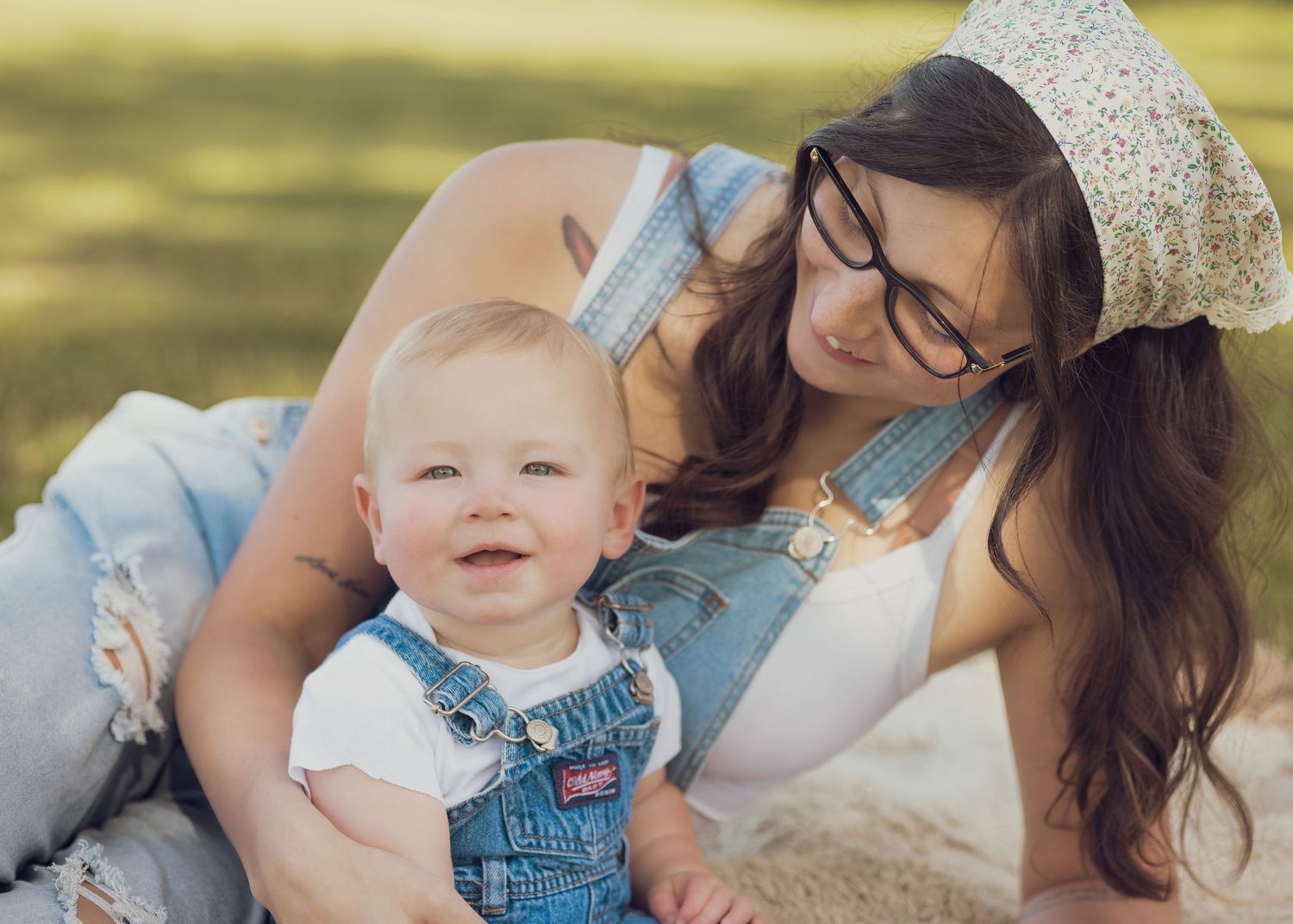 A young mother in denim overalls and floral hat sitting on a blanket with her happy baby outdoors.