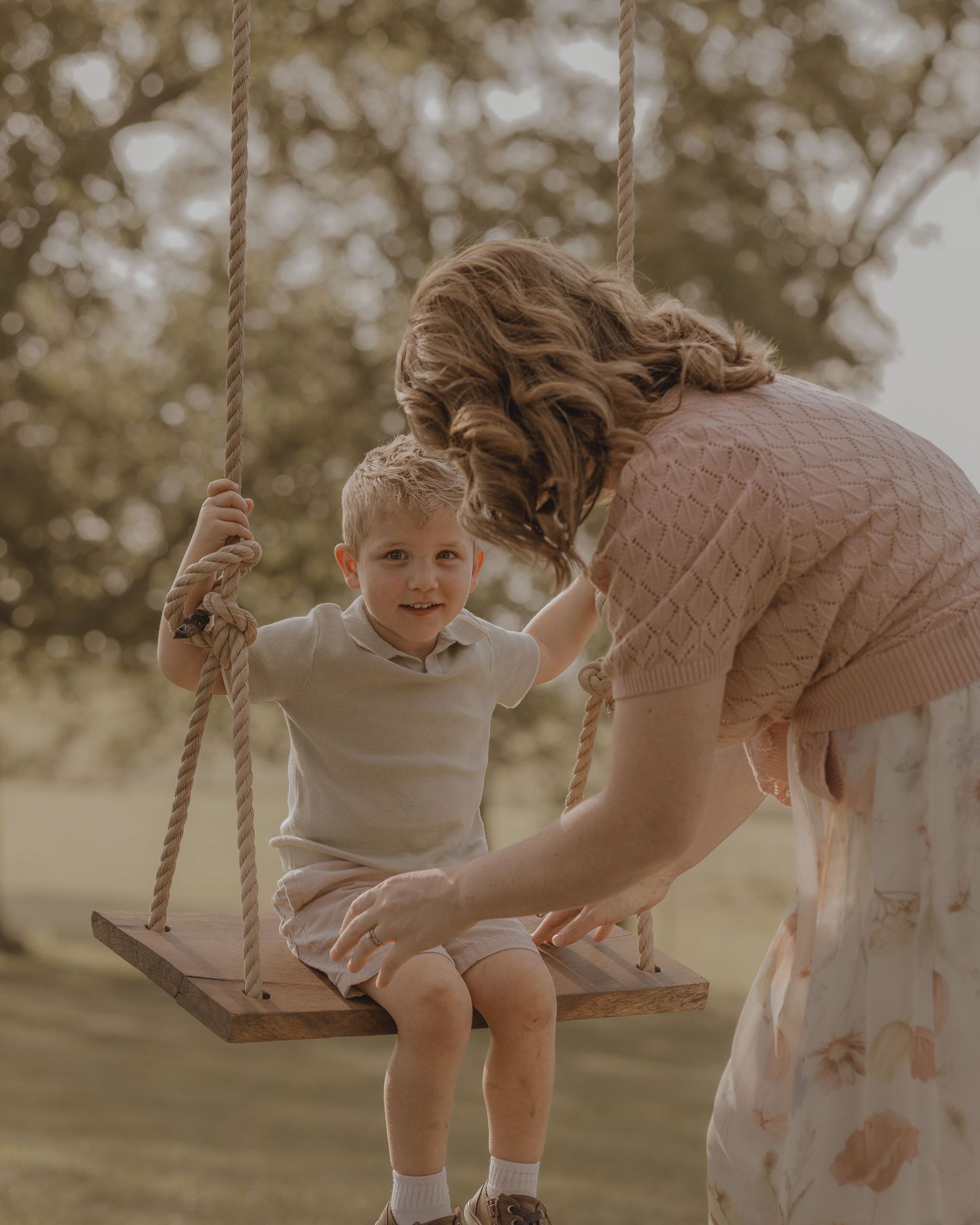 A mother gently steadies her toddler son sitting on a wooden rope swing outside.
