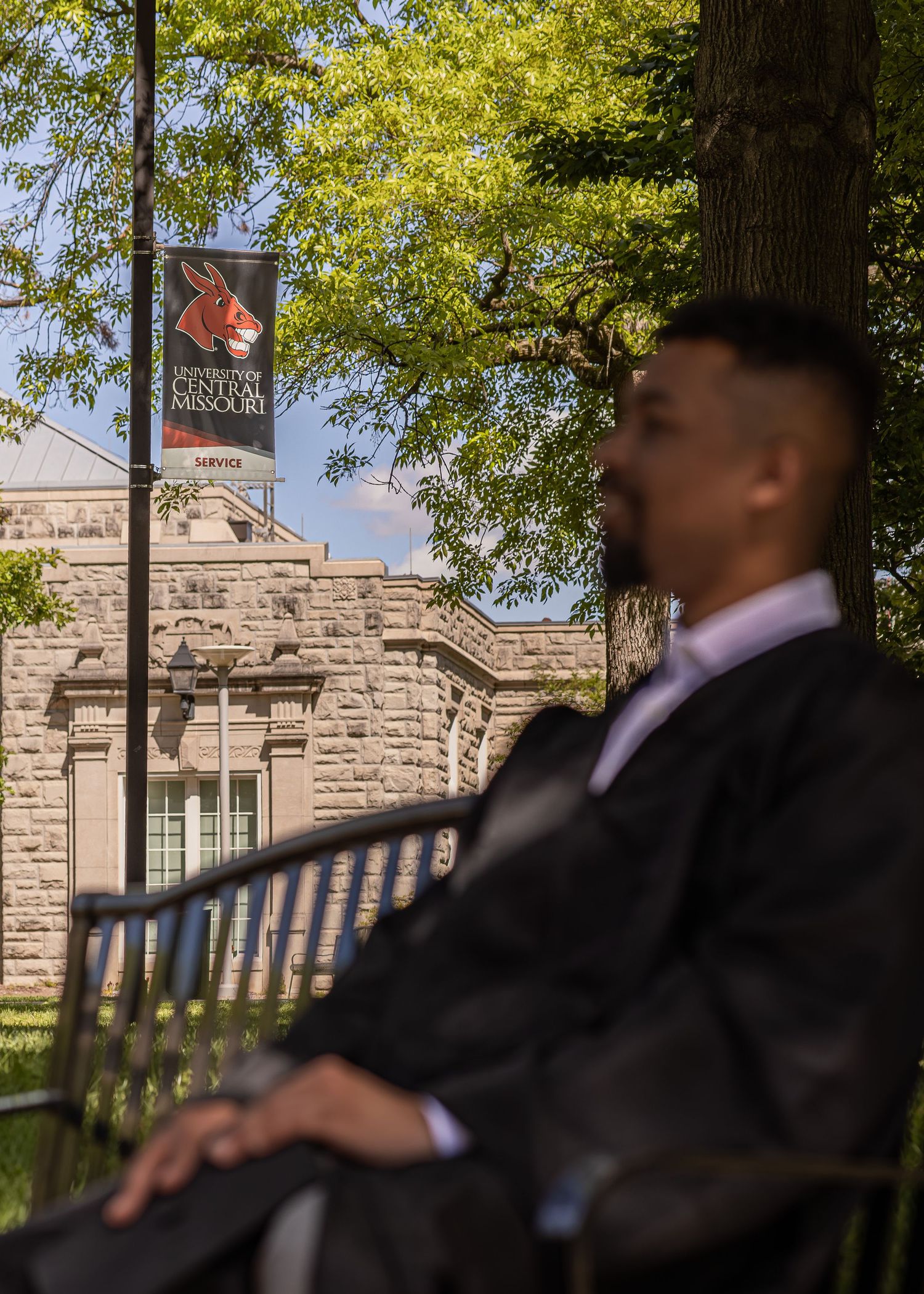 A graduate in cap and gown sits on a bench near a Central Missouri university banner and historic stone building.