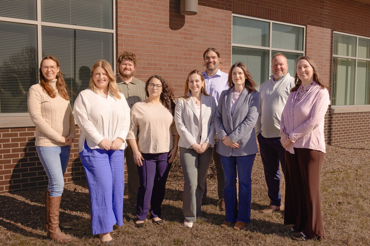 Group of nine professionals smiling outdoors in front of a brick building on a sunny day.