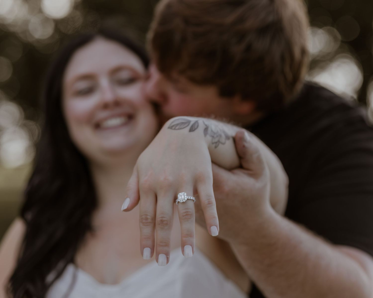 Couple celebrating engagement outdoors, woman smiling and showing diamond engagement ring on her hand while man kisses her cheek.