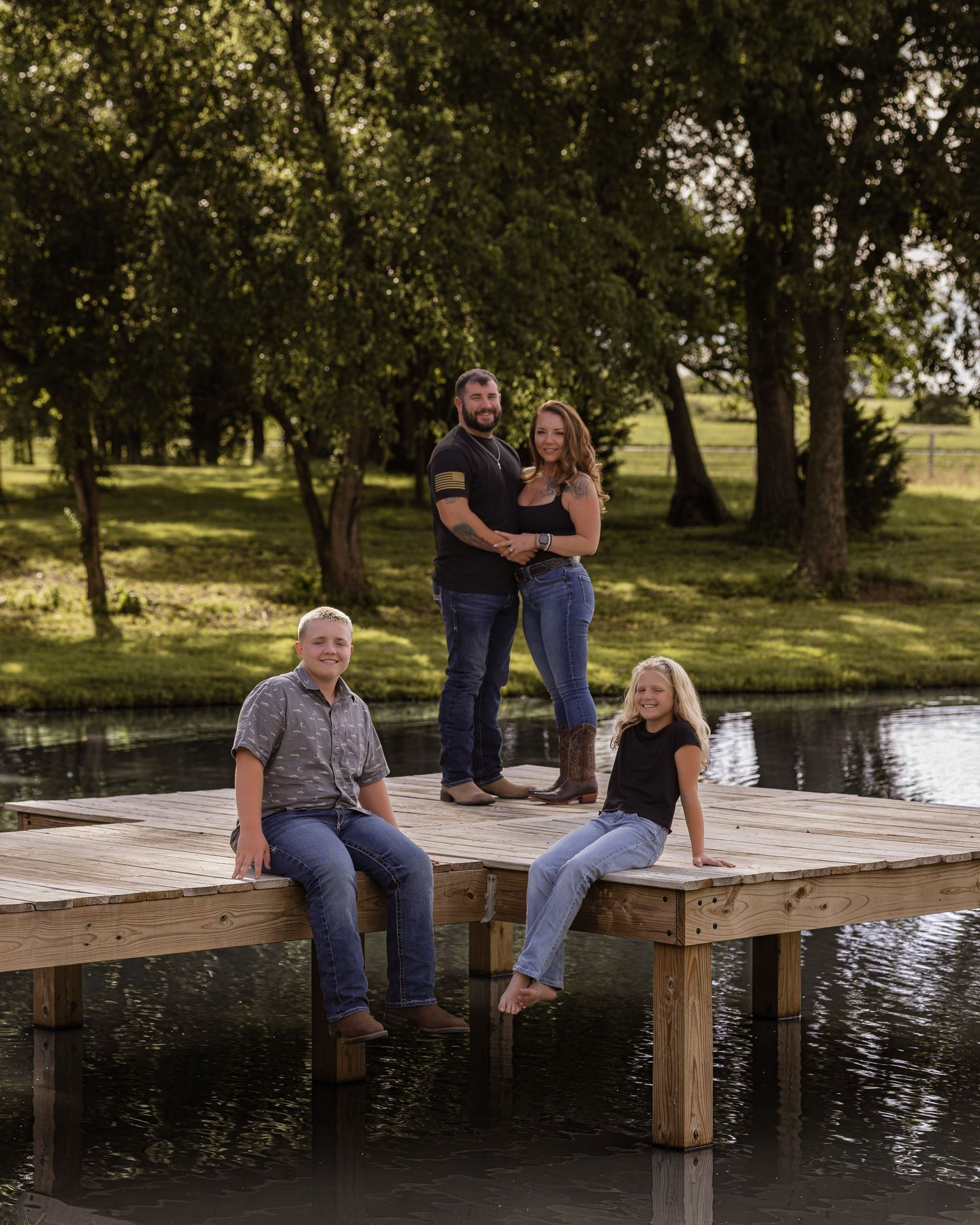 Family of four posing on a wooden dock by a pond surrounded by lush green trees on a sunny day.