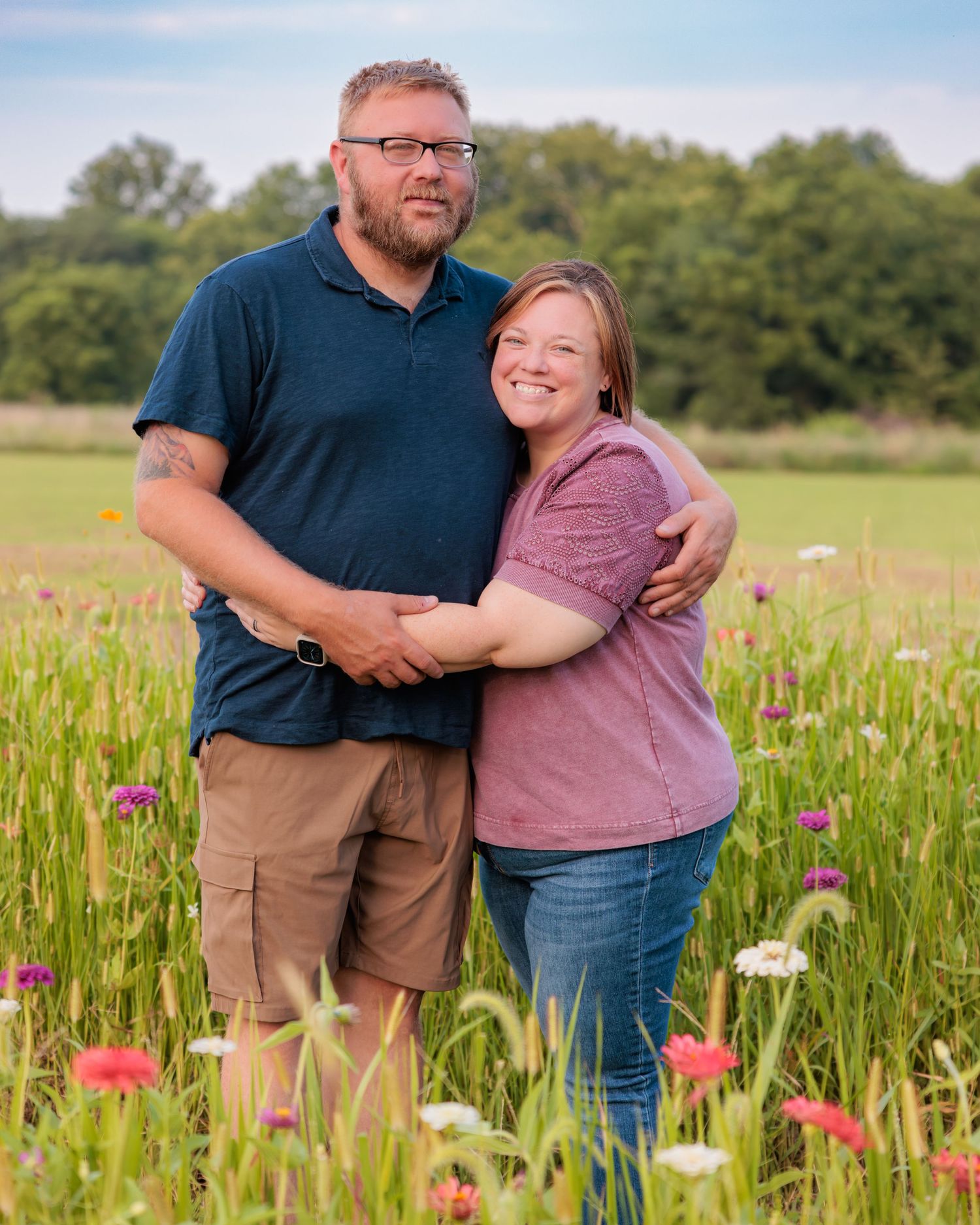 Couple embracing in a colorful wildflower meadow, smiling outdoors on a sunny day with lush green fields in the background.