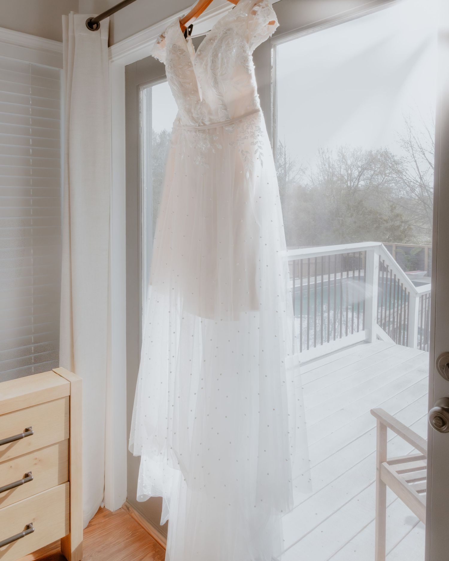 Elegant white lace wedding dress hanging on a curtain rod by a sunlit glass door and deck.