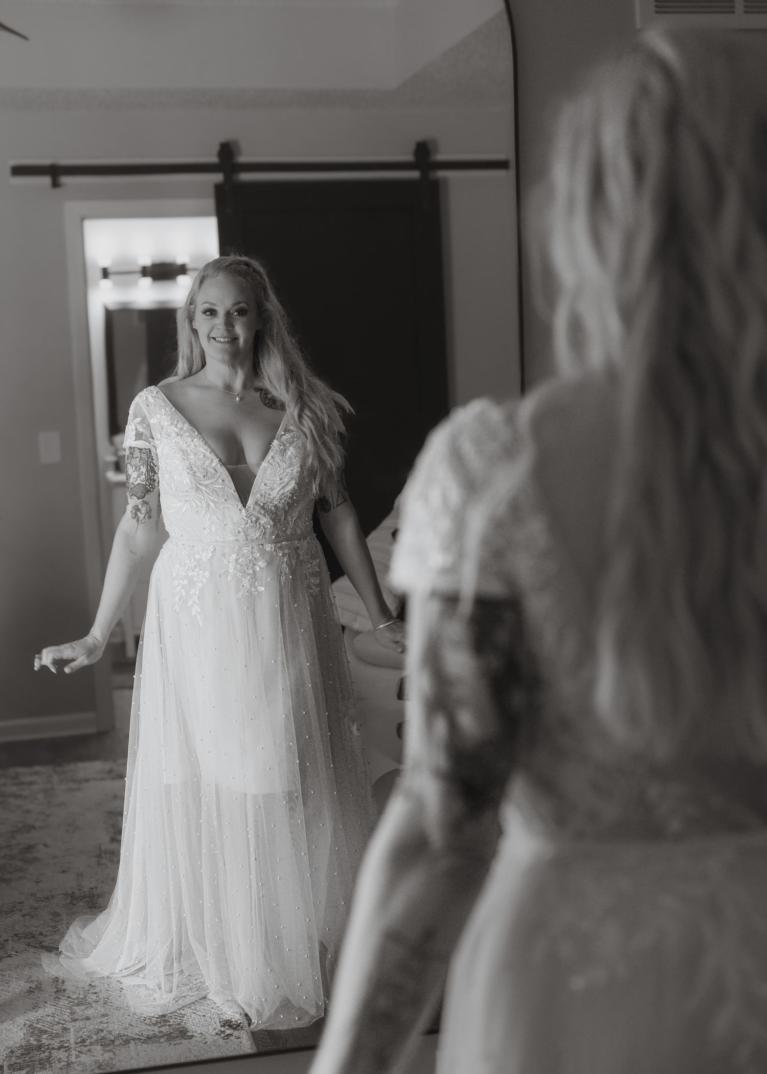 Bride in a lace wedding gown smiling at her reflection in a mirror during wedding preparations.