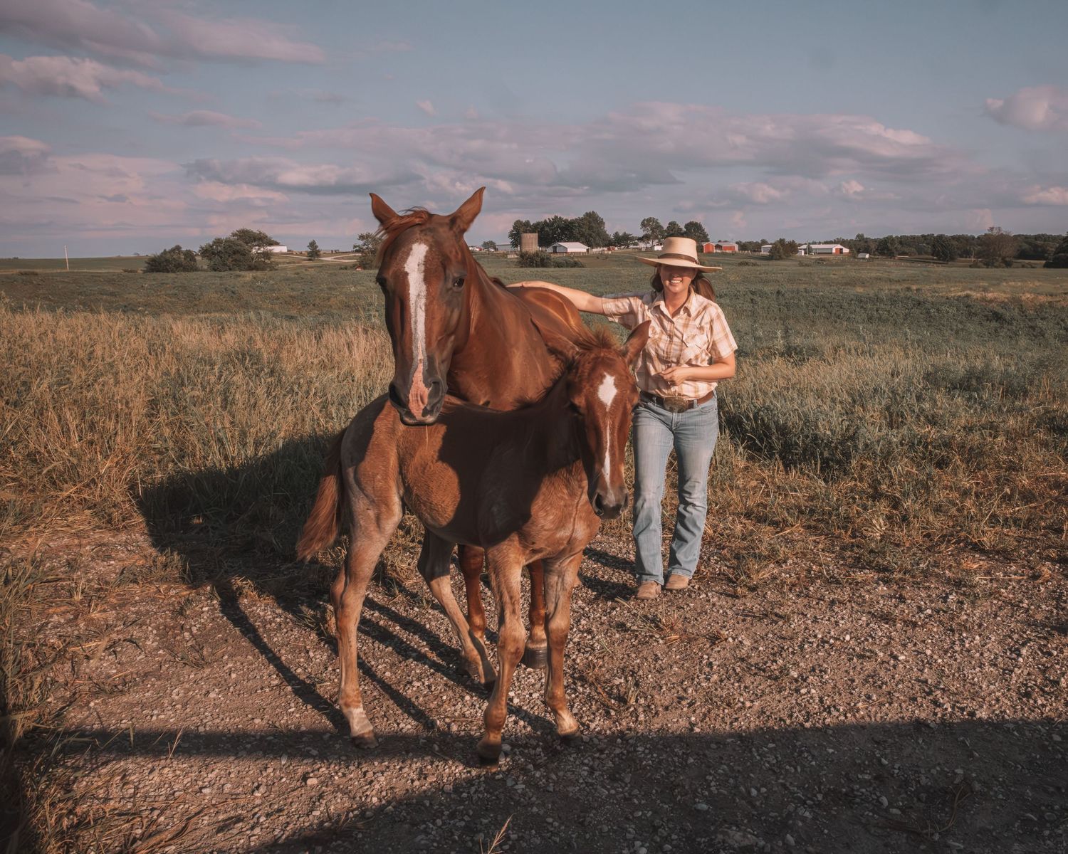 A cowgirl stands with a chestnut mare and foal on a rural farm dirt path at golden hour.