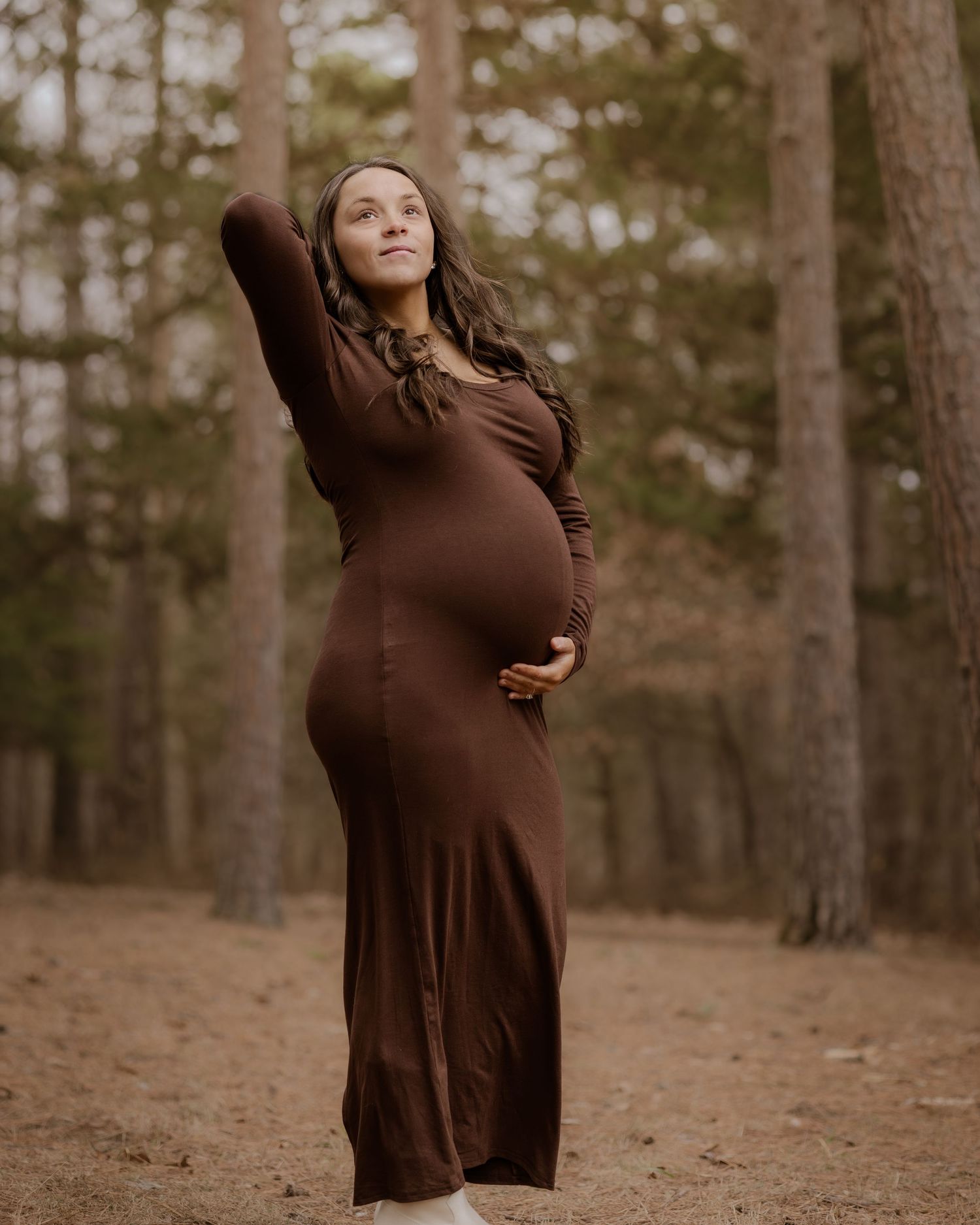 A pregnant woman in a brown dress poses gracefully in a wooded forest setting during a maternity photoshoot.
